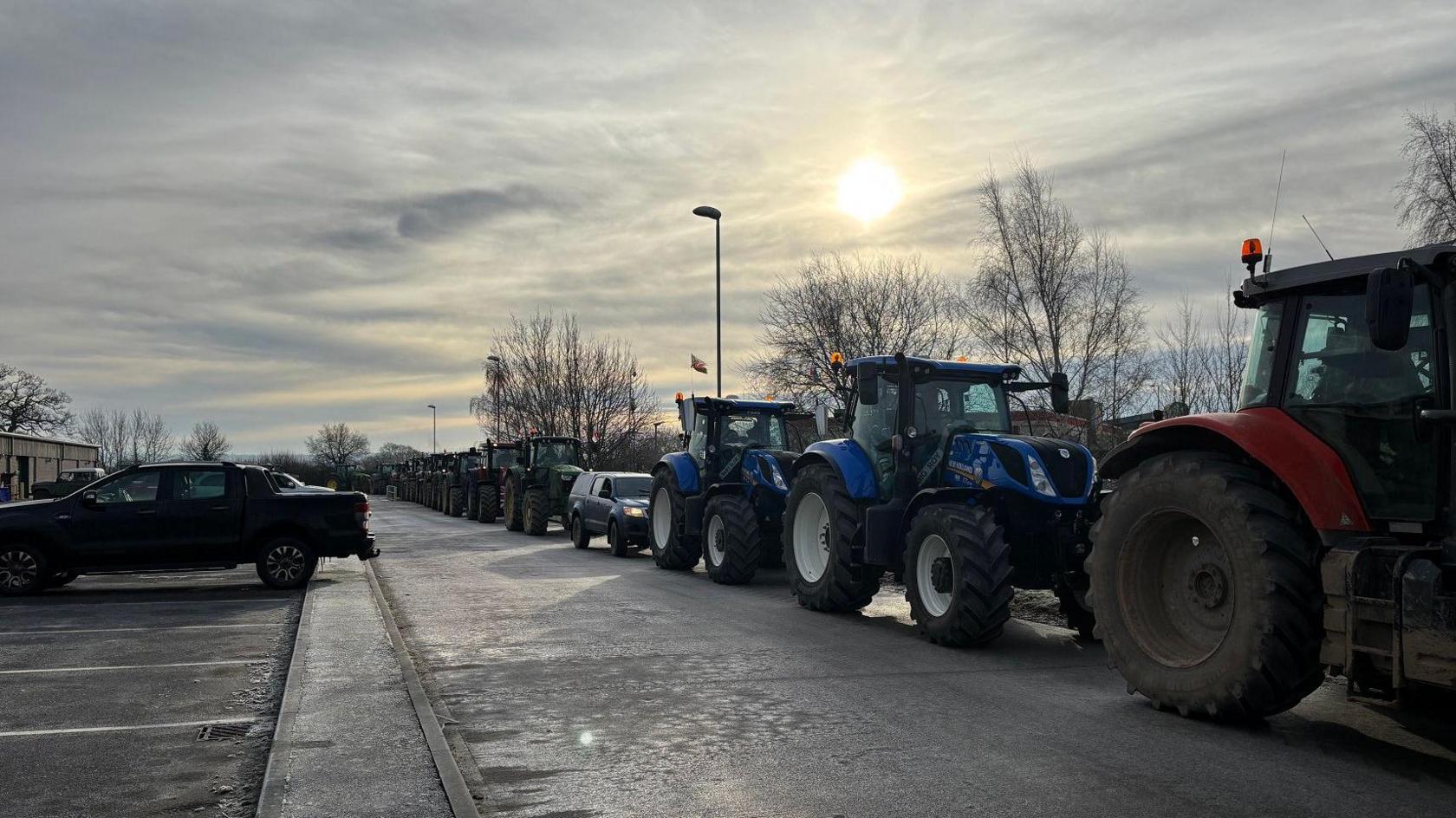 A long line of tractors are on a road. They are different colours, including red, blue and green. The sky is bright white white and cloudy above them.