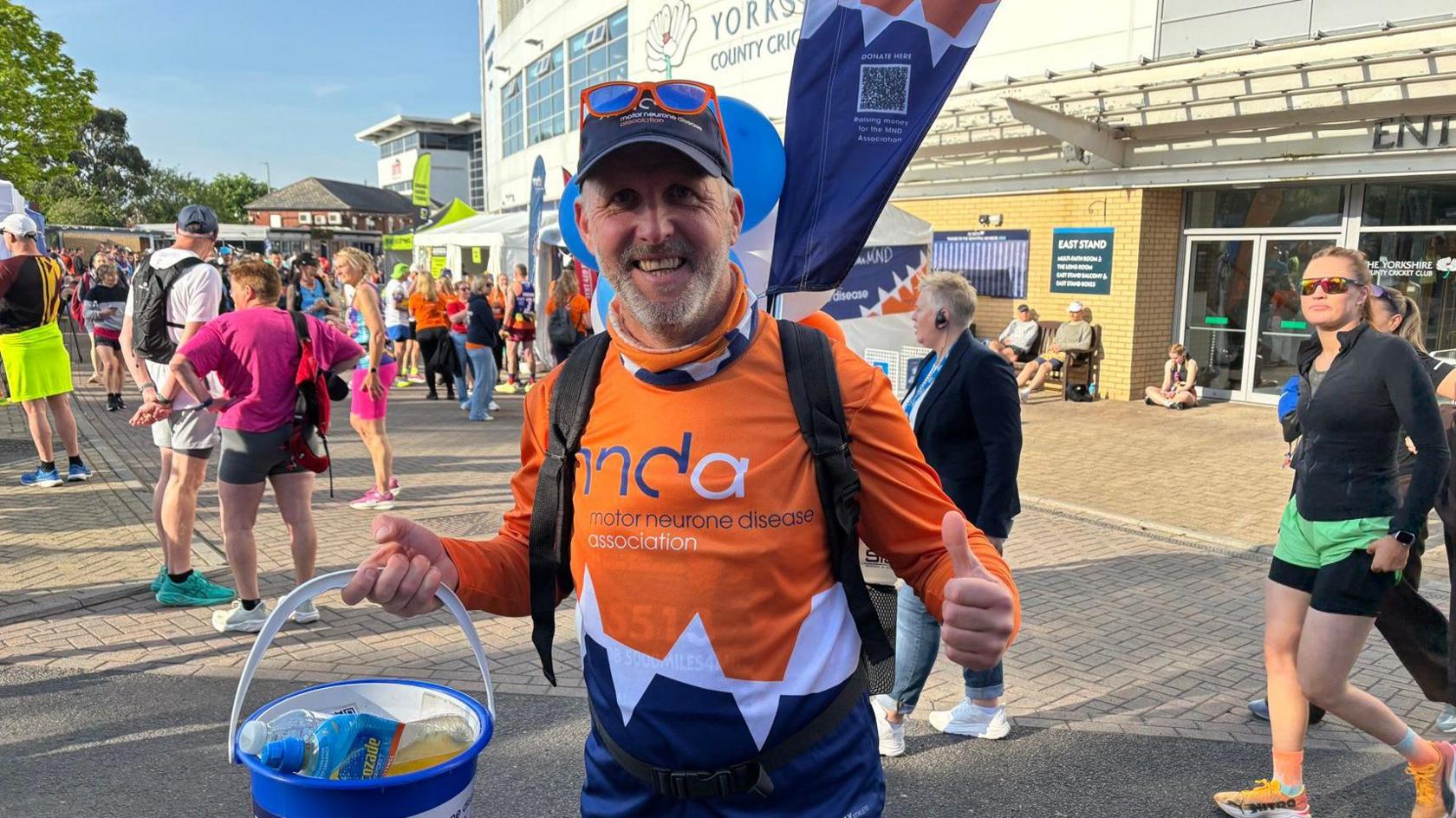 A man in an orange MND association t shirt. He has a flag on his back and a collection bucket which, today, contains bottles of water instead of coins
