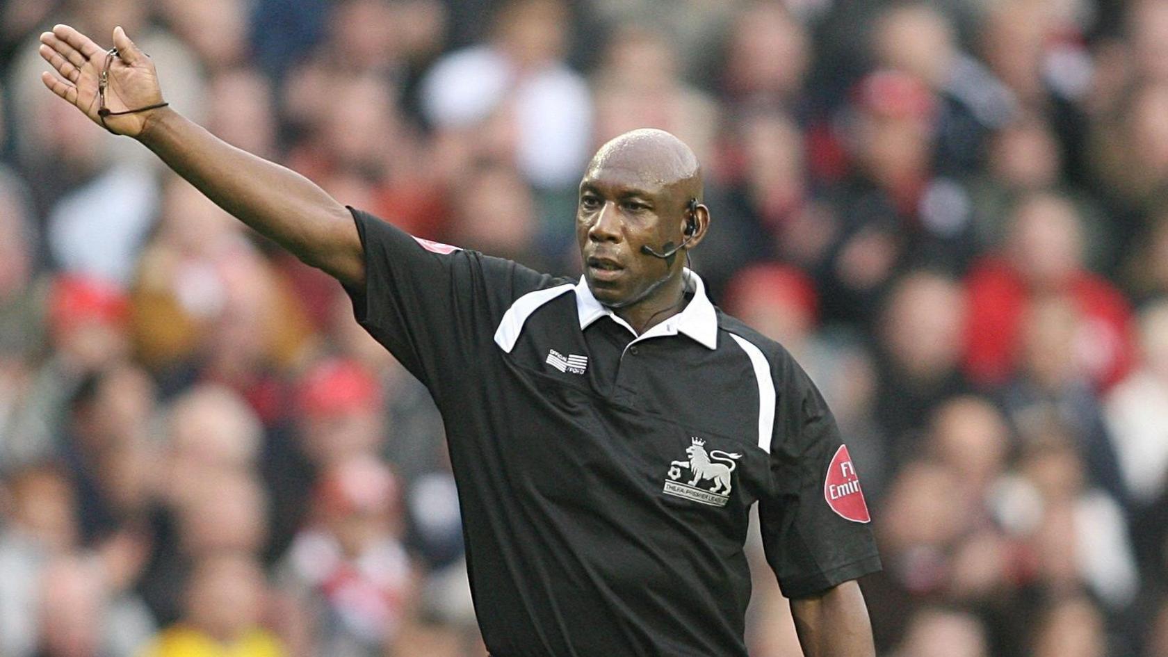 A man wearing a black referee's top with a Premier League logo on the chest. He is wearing a microphone and earpiece. He has one arm outstretched. In the background is a blurred view of fans in the stadium.