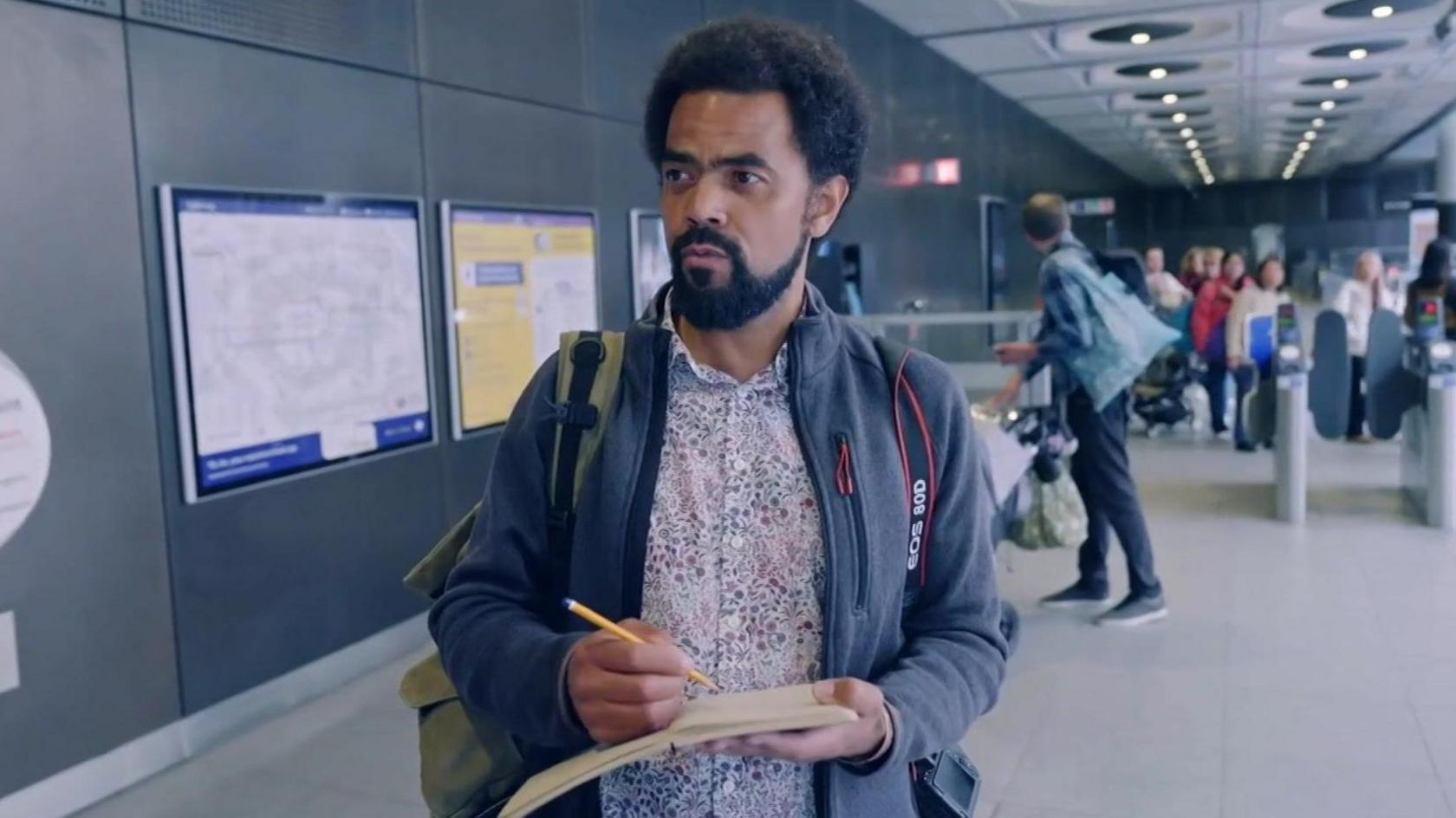 Jack stands in a busy train station, holding a notebook and pencil, appearing focused. He wears a patterned shirt, a grey jacket, and carries a backpack, with a camera strap over his shoulder.