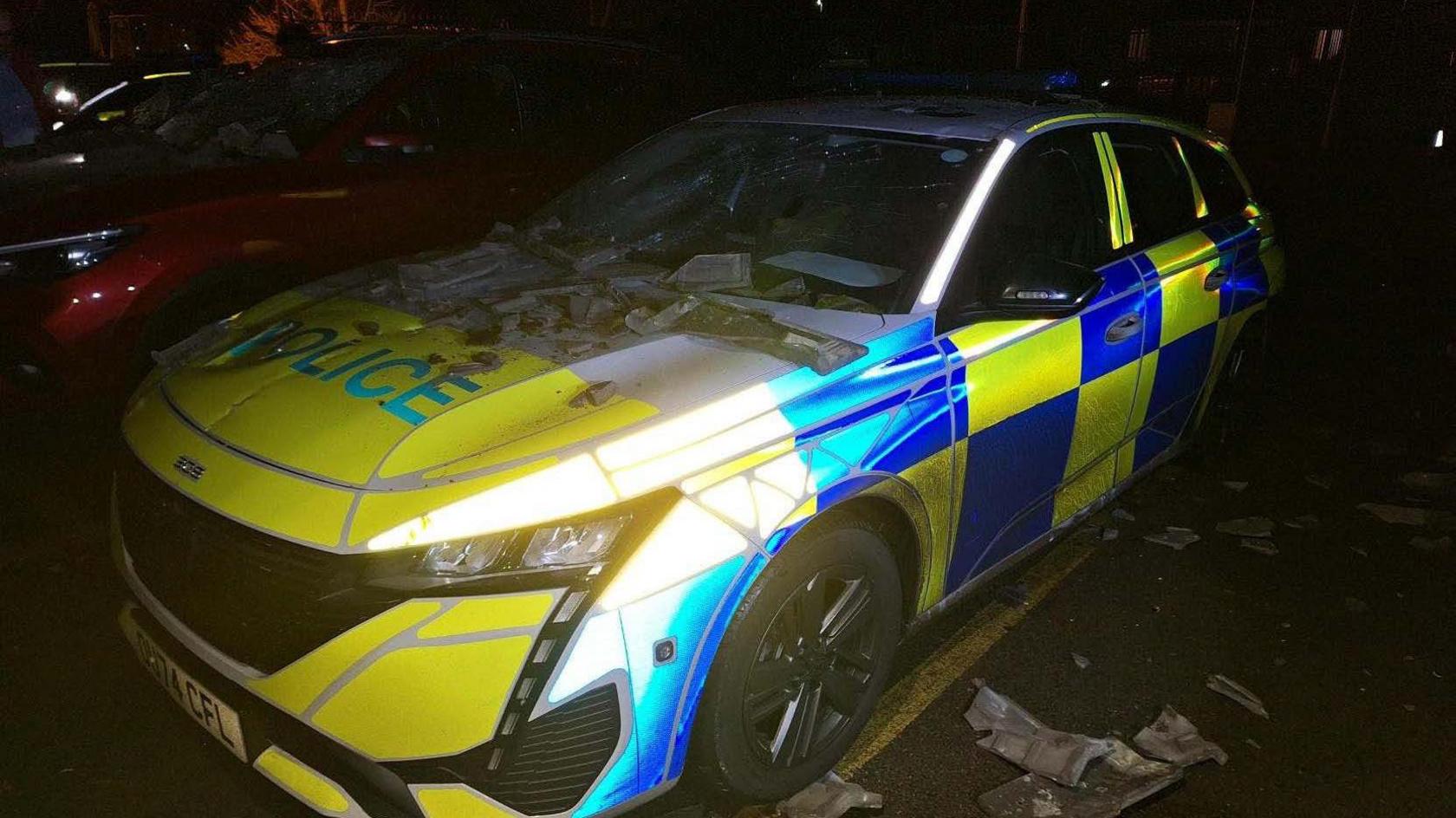 Another view of the police car from the side with smashed ceramic tiles on its front windscreen and bonnet. The windscreen has been smashed by the tiles. Smashed tiles can be seen on the ground around the vehicle.