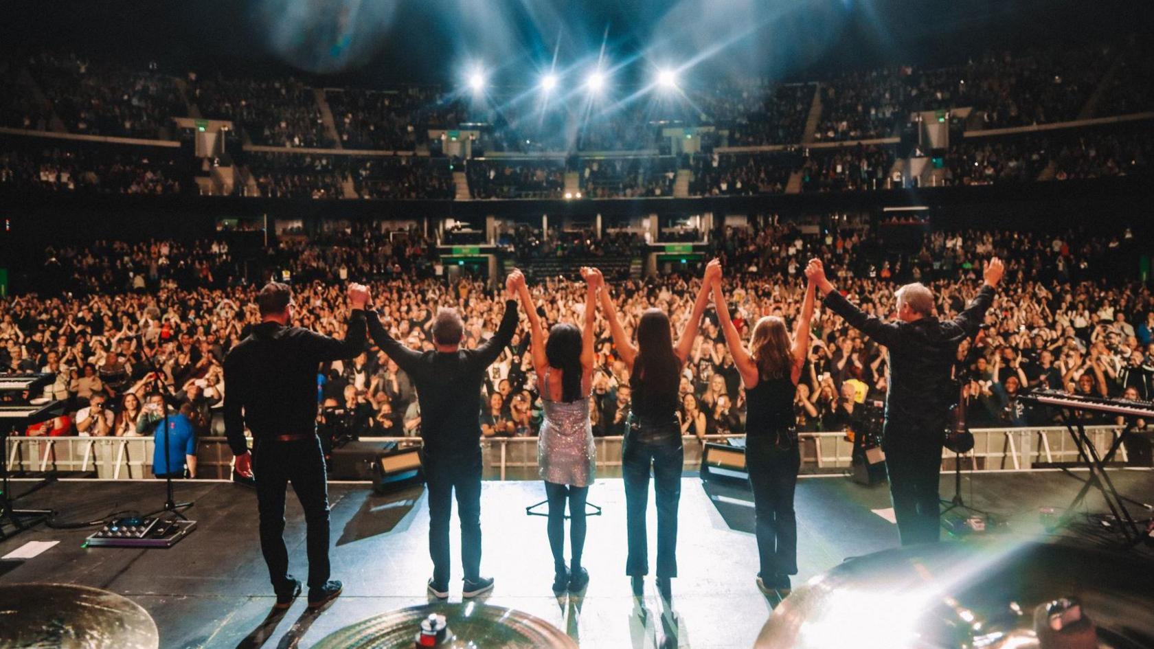 Éadaoin Ní Mhaicín is on stage with The Corrs. They are seen from the back, facing the huge crowd in the auditorium. Six people stand in a row, Éadaoin Ní Mhaicín is third from right. They are holding their hands in the air, reacting to the applause.