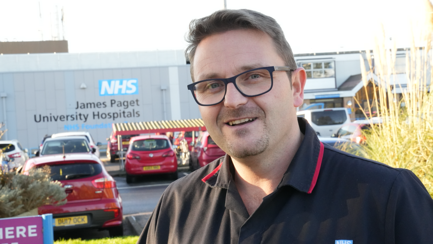 A man standing in front of a hospital. He is wearing glasses and a black polo-shirt. He is smiling at the camera.