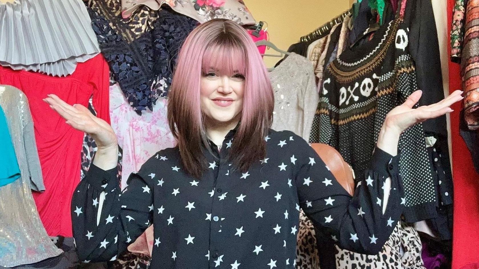 Caroline in her spare room, full of charity shop items. She is holing her arms out, wearing a pink and brown wig, with a black top with stars on, with clothing all around her. 