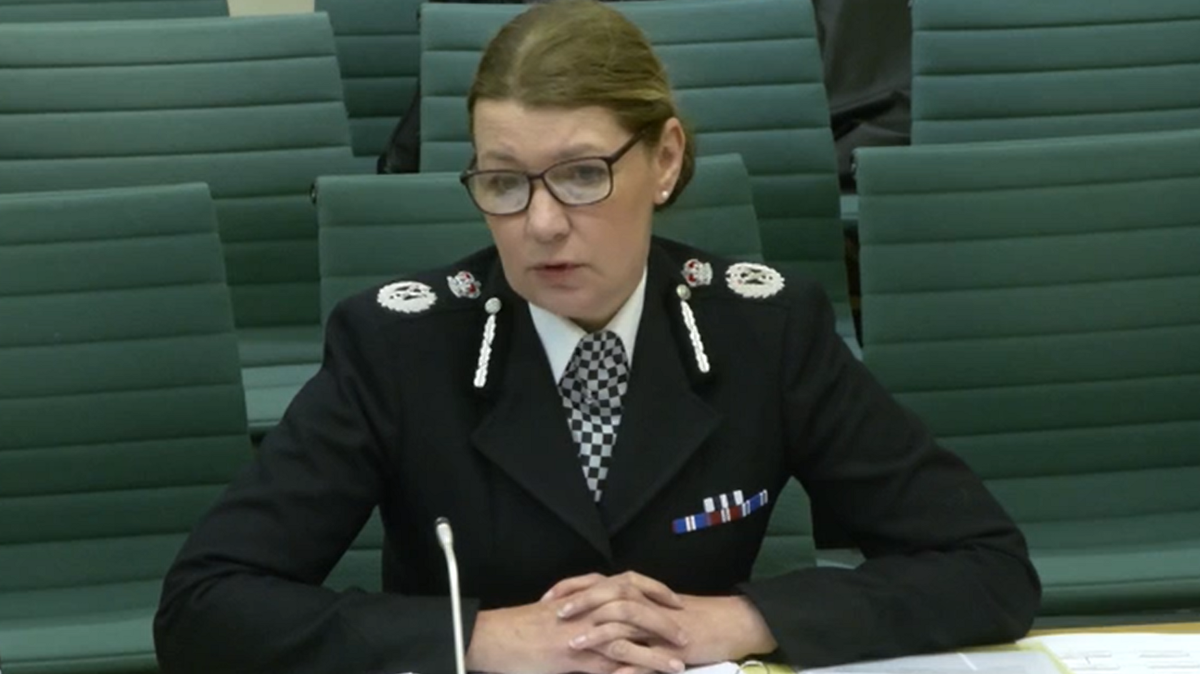 Judi Heaton is sitting behind a desk with her hands clasped together. She is wearing a traditional police uniform with her silver coloured badges clearly visible against her black jacket. She is wearing glasses and her brown coloured hair is being worn up.