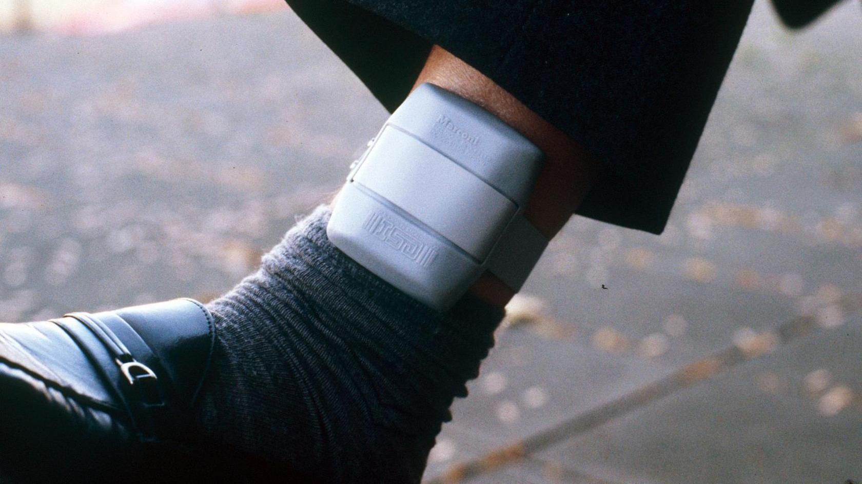 A close-up image of a grey ankle monitor. It is above a dark grey sock and black shoe on someone's leg