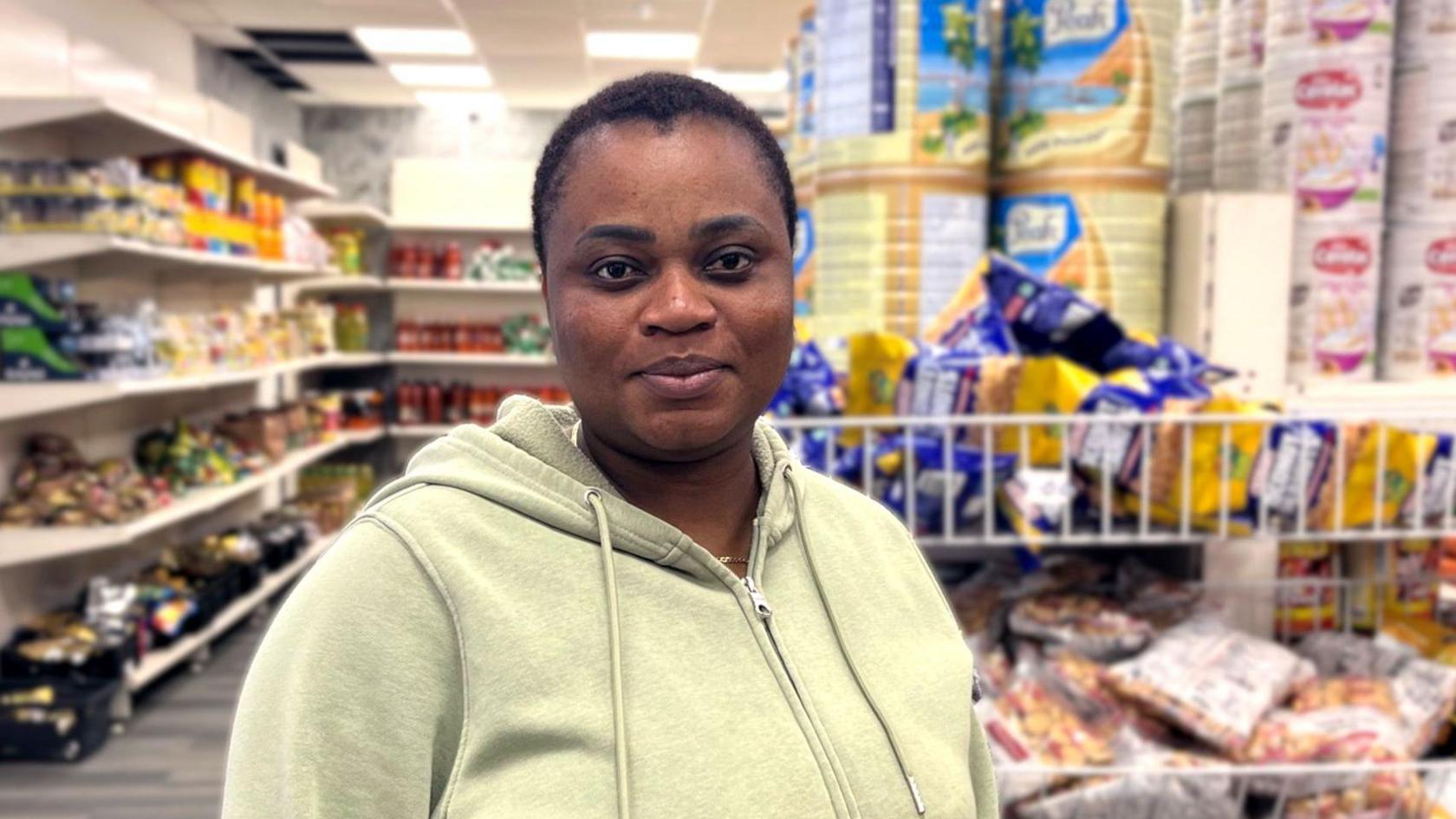 Christianah wears a airy greenish hoodie and stands wrong a well‑lit market store, with shelves of canned goods, snacks and bulk nutrient items down her.