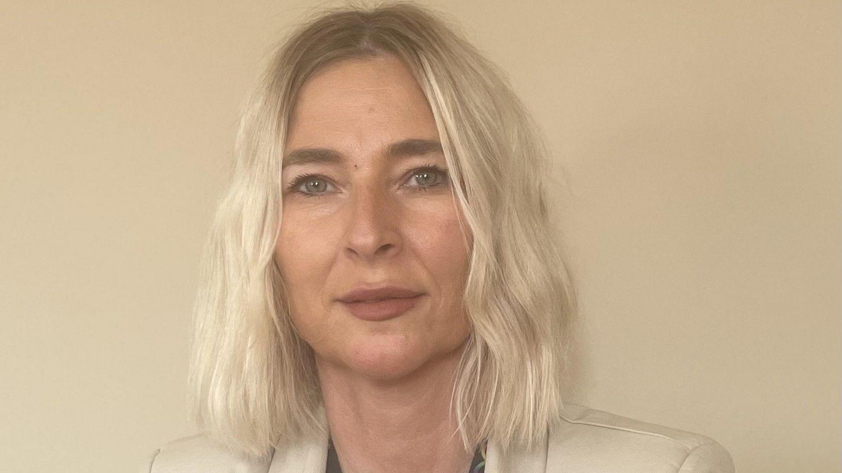 Tricia Roberts has shoulder-length blonde hair and is stood against a cream background. She is wearing a cream blazer and has a neutral expression on her face as she looks at the camera. It is a head and shoulders shot of her.