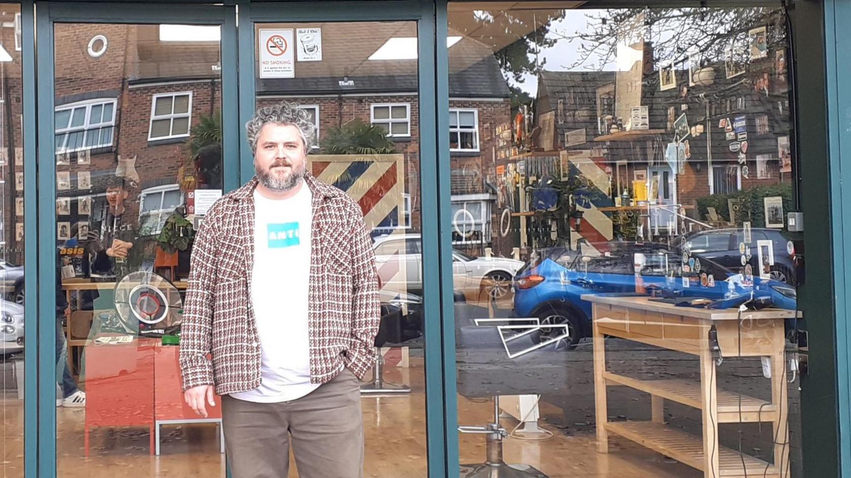 A man in an open chequered shirt standing in front of his barber shop frontage