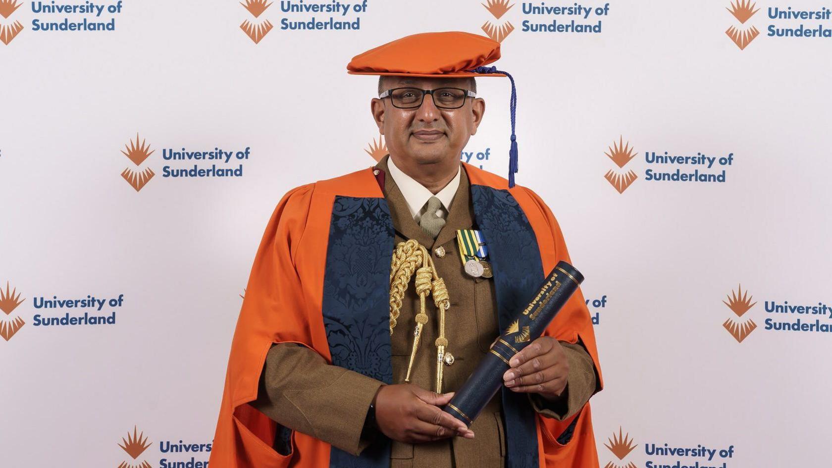 Ben Banerjee receiving his honorary doctorate of medicine from the University of Sunderland. He is wearing an orange academic cap and gown as well as a brown military uniform. He is holding a scroll and looking into the camera.