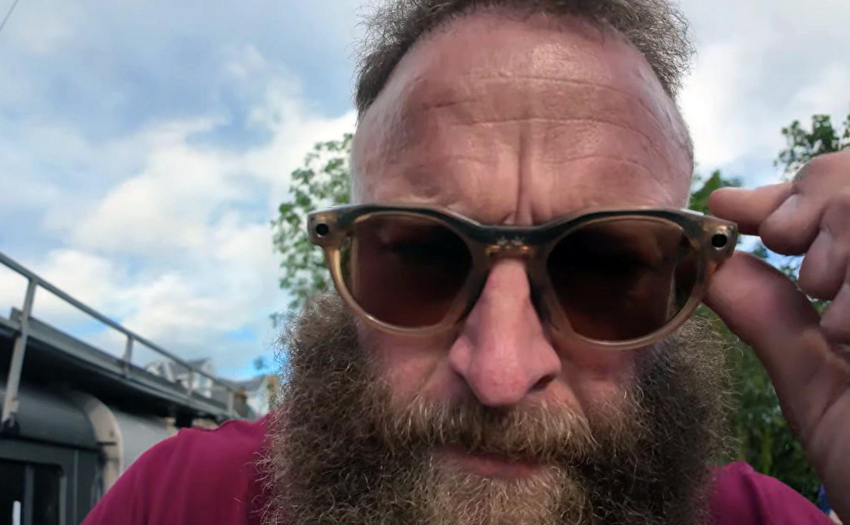 A bearded man in a red T shirt wearing sunglasses looks at the camera