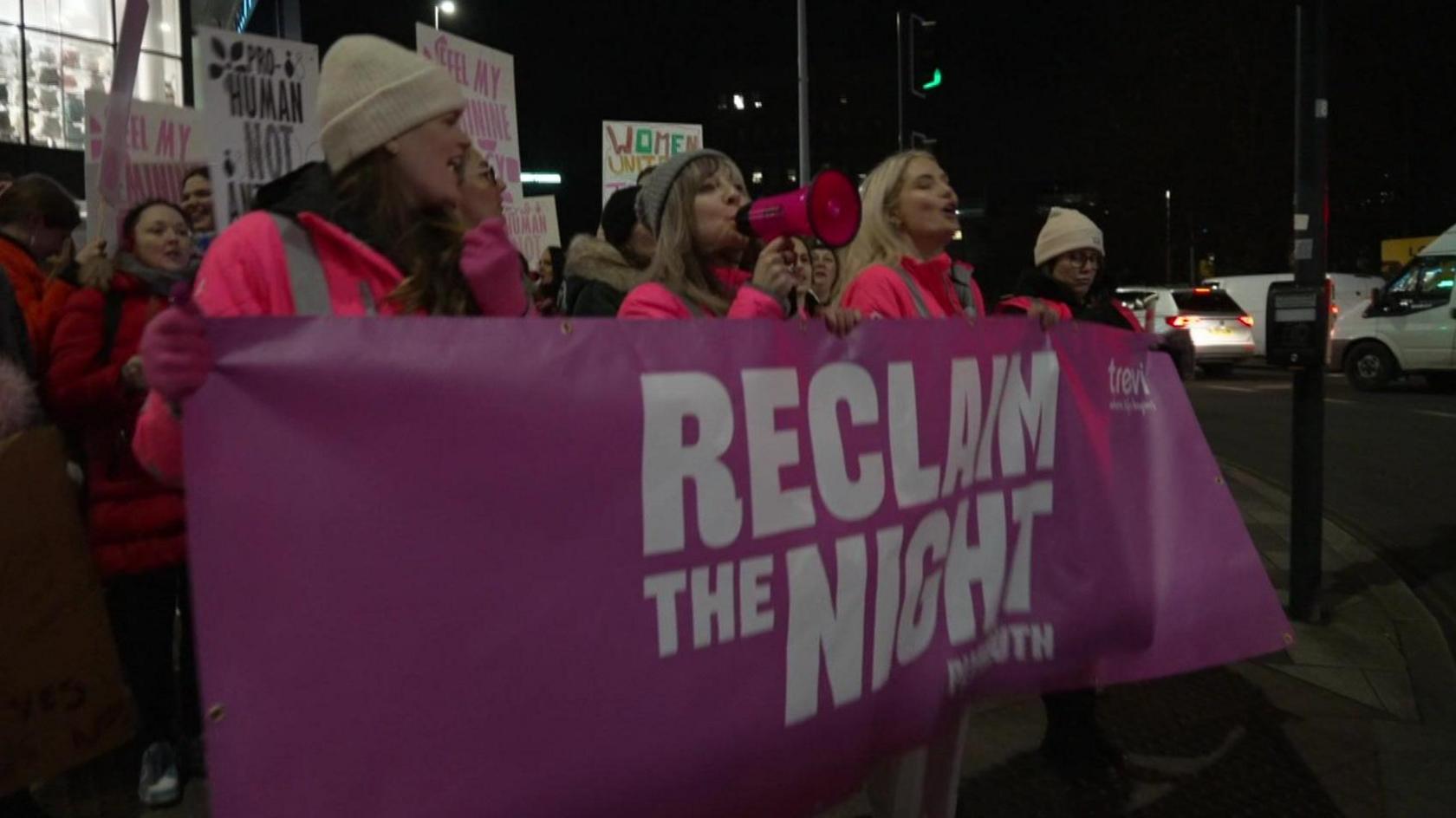 People in Plymouth city centre taking part in a Reclaim the Night march. There is a group of women wearing bright pink high vis jackets holding a pink banner which says reclaim the night Plymouth.