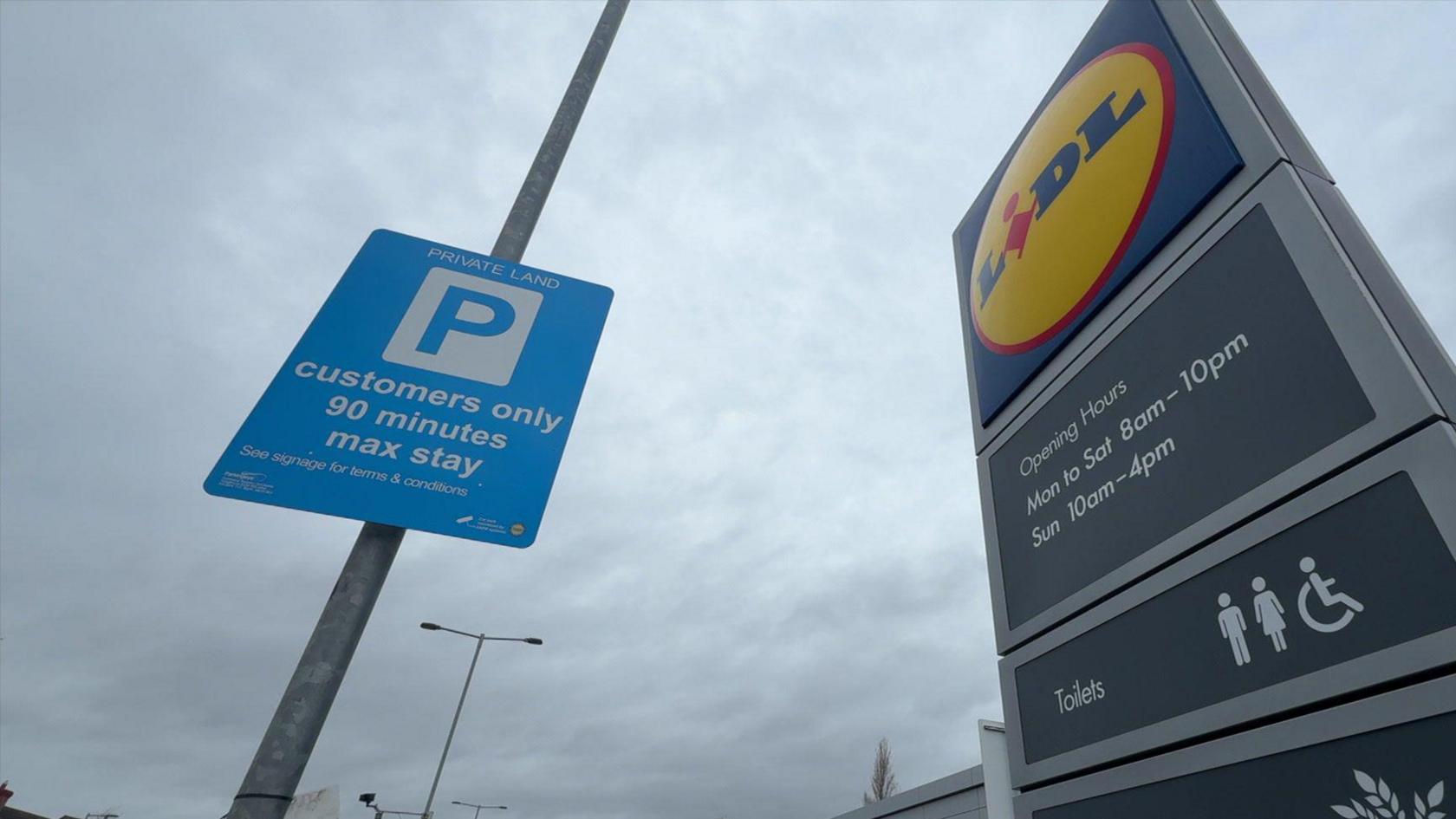 Two signs in a supermarket car park. A blue rectangular sign on the left of the image has been fixed to a lamp post and reads "Private land, customers only, 90 minutes max stay". Another sign on the right of the image features a blue, yellow and red Lidl logo and has details of the store opening hours. Both signs are set against a cloudy background.