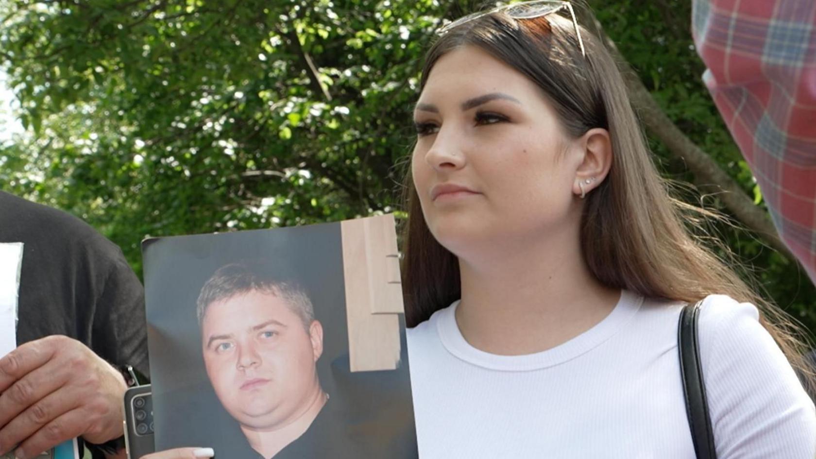 A woman in a white T-shirt and brown hair holds a photo of a young man