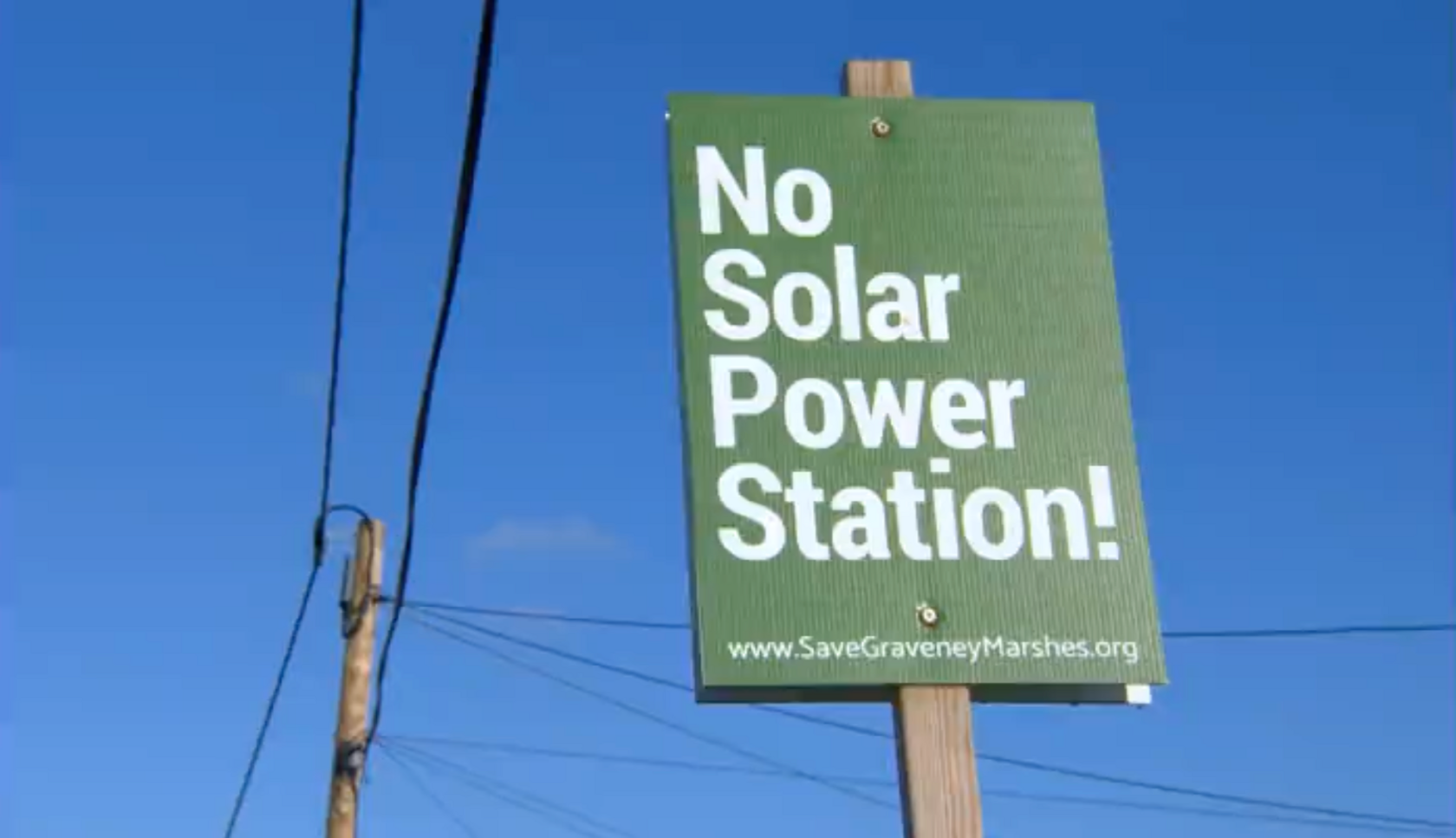 Green sign saying "No Solar Power Station!" on a placard photographed from below showing the sky and power lines behind it