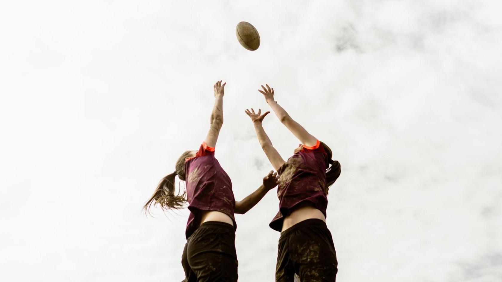 Two girls reaching for a rugby ball in the air. The photo is against a cloudy sky.