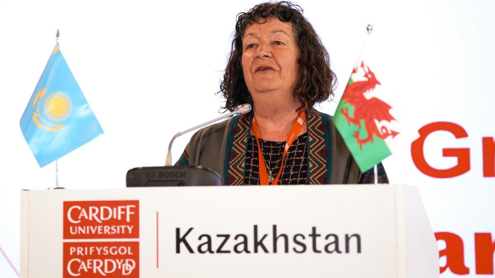 Menyw gyda gwallt brown yn sefyll tu ôl i ddarllenfa sy'n dweud Prifysgol Caerdydd Kazakhstan a baneri Kazakhstan a Chymru arno