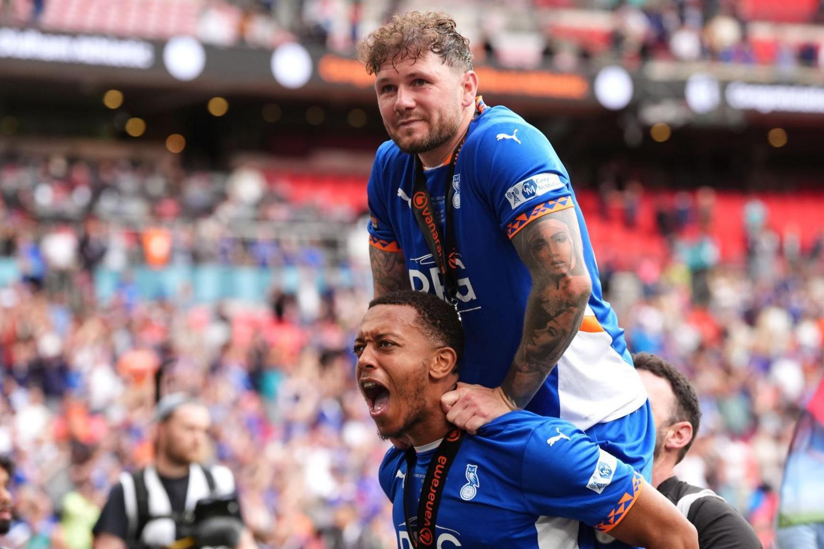 Shaun Hobson wearing an Oldham Athletic jersey and a medal round his neck. He appears to be shouting, with teammate James Norwood leaning on his shoulders. In the background are the crowds in Wembley Stadium, but out of focus.