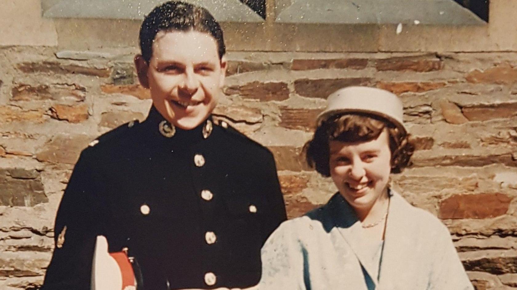 Mr Latham wearing a dark blue military uniform with gold buttons next to his wife Evelyn, who is wearing a light blue coat with a matching hat. 