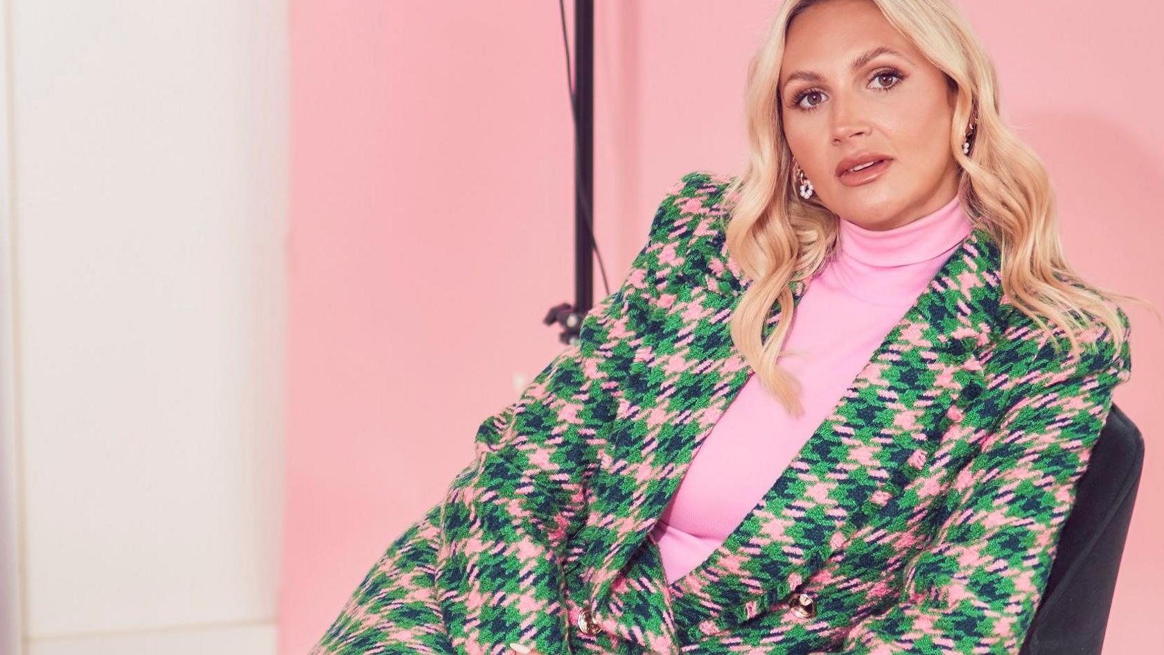 Sophie Pycroft looking at the camera on a chair, sitting slightly sideways. She is wearing a checked skirt suit in green and pink and a pink top.