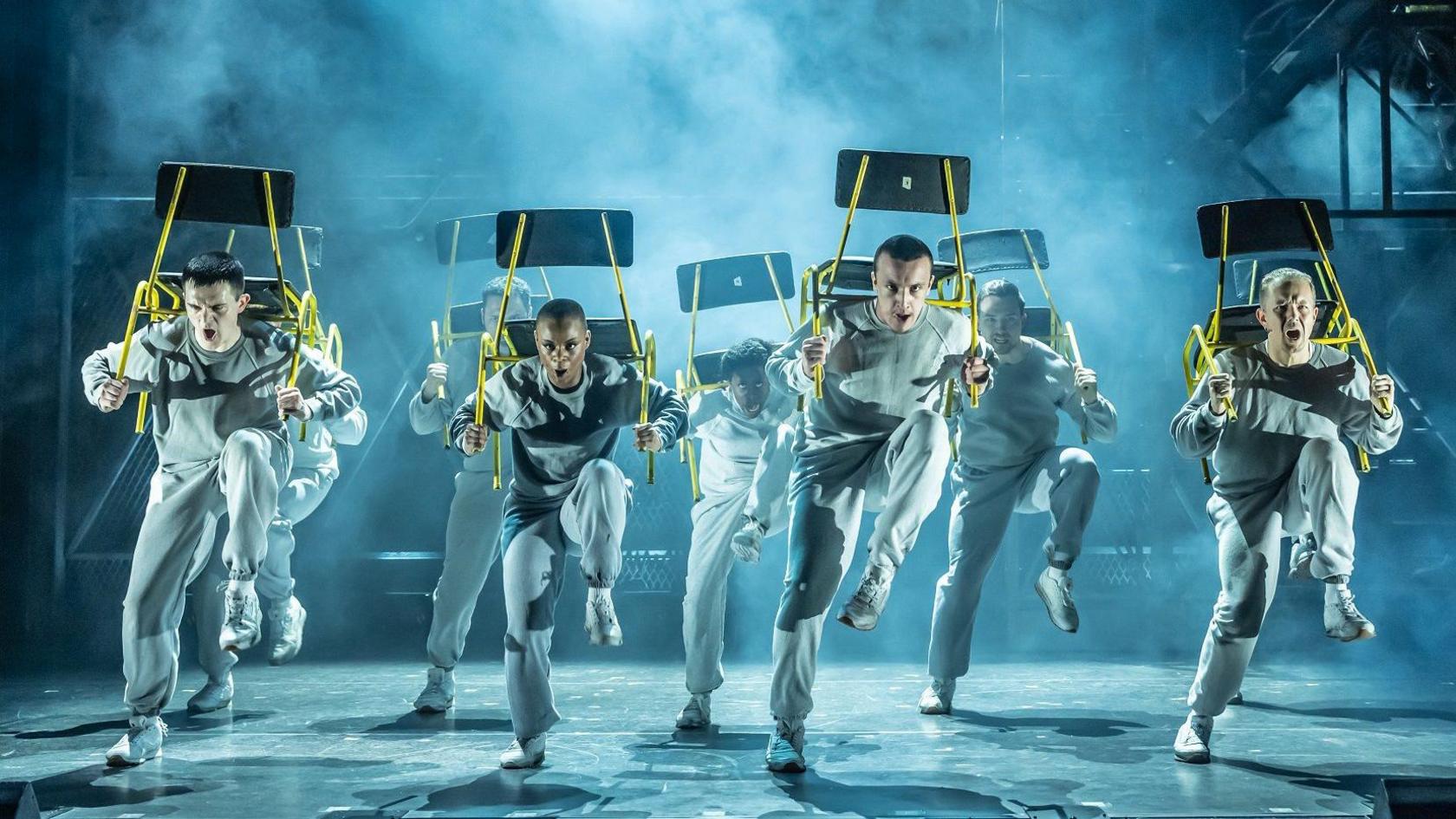 A group of performers in white outfits dance in formation during a production of an Officer and a Gentleman at the Bristol Hippodrome. They are looking stern and carrying chairs on their backs