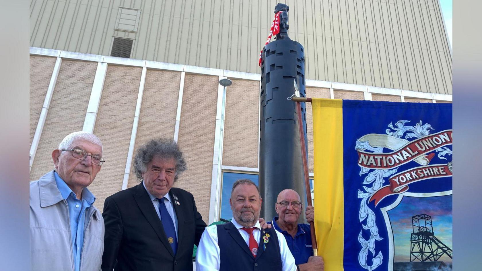 Four men around their sixties, dressed smartly, stood infront of a black column statue with a child at the top