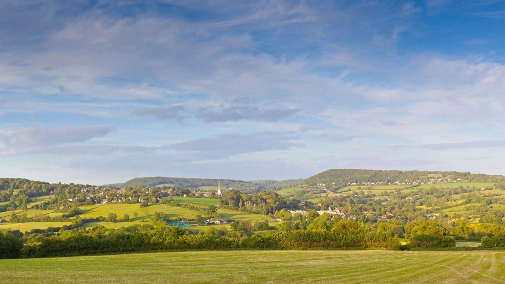 A wide vista of a typical Cotswold landscape of rolling hills and woodland copses. A big blue sky with whispy cirrus clouds.