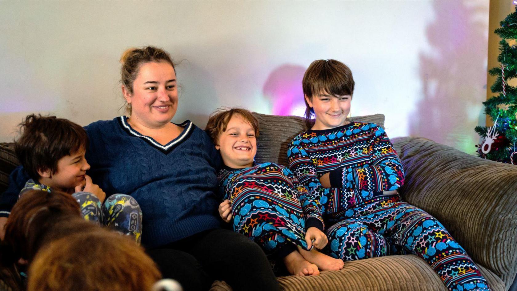 Sammiie sitting on the sofa wearing a blue knitted jumper with her dark blond hair tied up into a bun. She has her arms around her kids, who are sitting beside her wearing blue Christmas pyjamas. Next to the sofa is a Christmas tree decorated with tinsel, coloured lights, a reindeer and a candy cane ornament. All of them are lit up by the TV in front of them. 