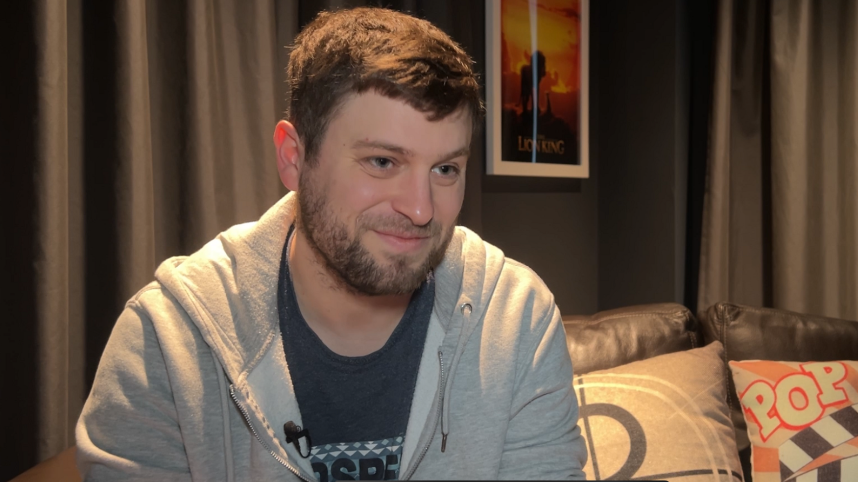 Joshua is sat on a leather sofa, with cushions behind him. On the wall behind him are cinema posters. He is wearing a grey hoodie, blue t-shirt and has short brown hair. 