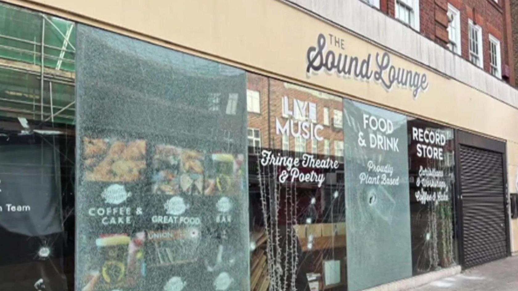 A shop exterior, made of glass. Each panel is smashed or damaged in some way.