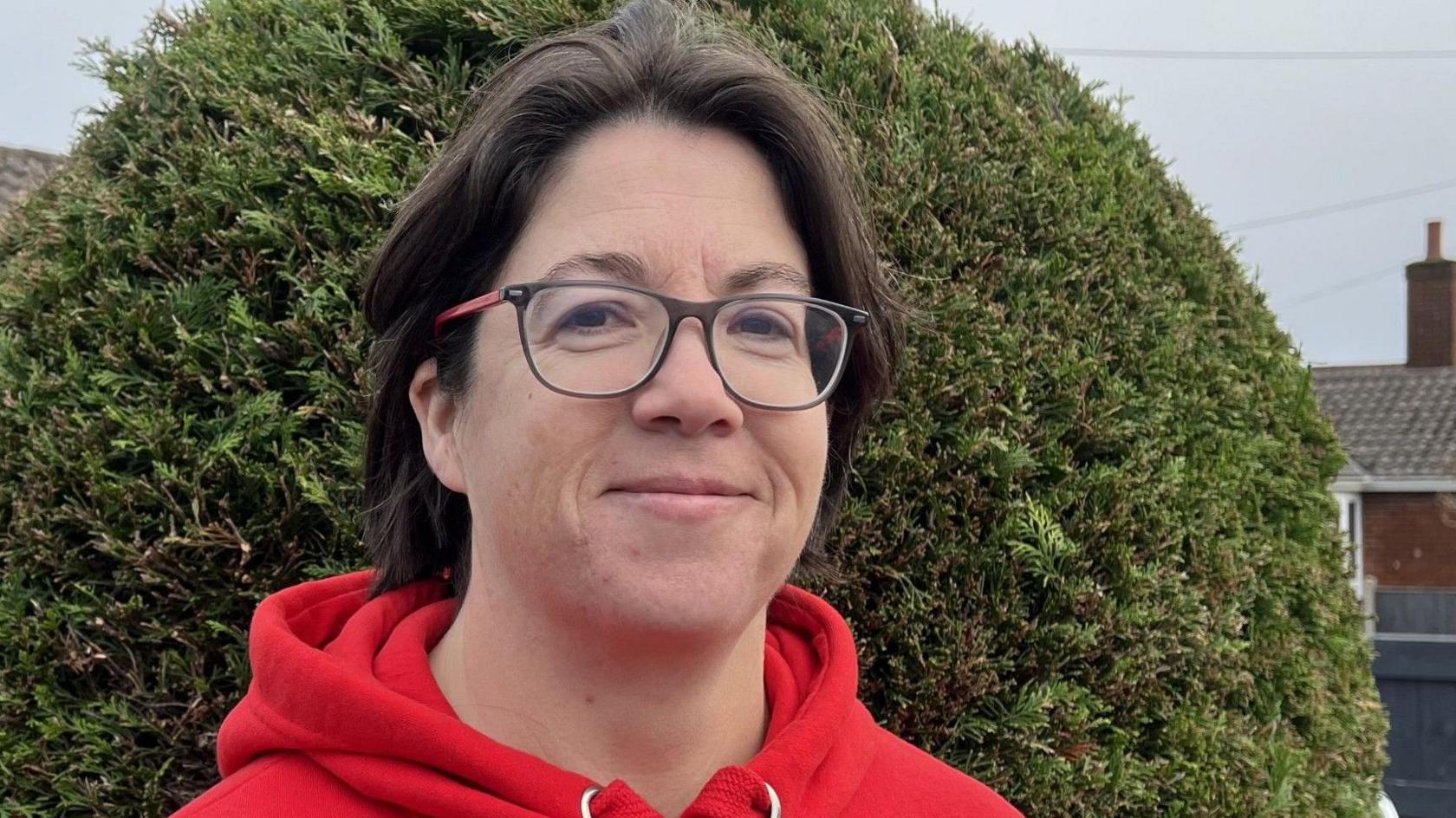 Woman with short dark drown hair and wearing a red hoody and black and red framed glasses smiling, standing in front of a dense bush. 