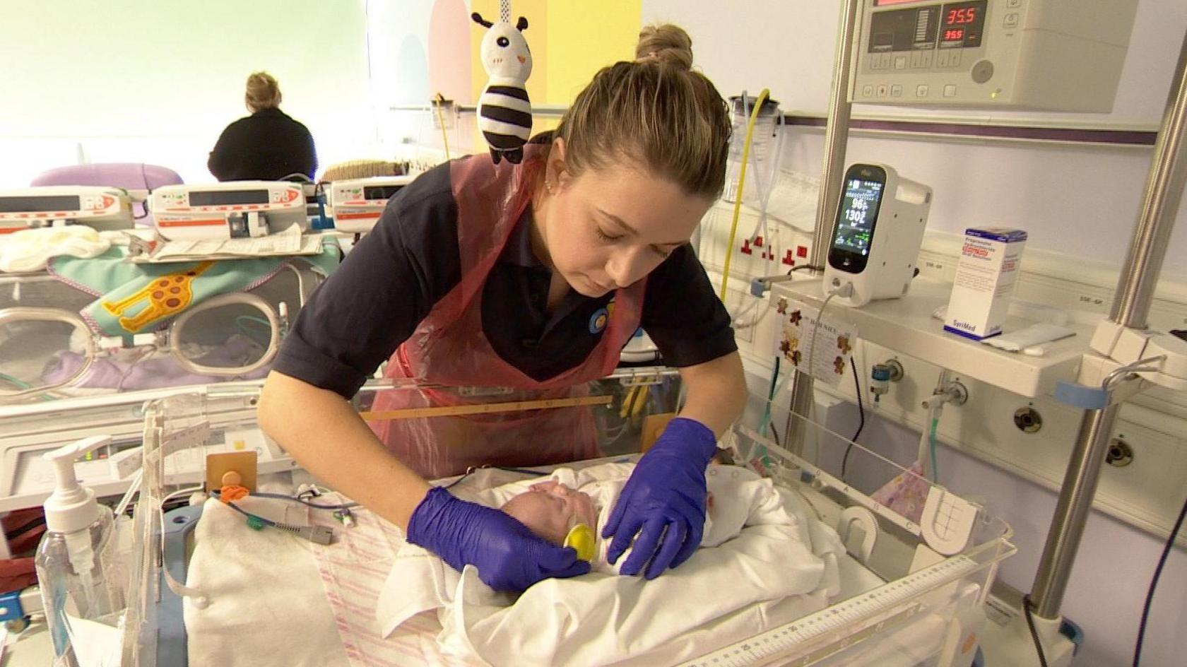 A female specialist neonatal paramedic, who is wearing a black shirt and  disposable red apron, fits yellow foam cups on to the ears of a tiny baby who is laid in a cot surrounded by medical equipment.