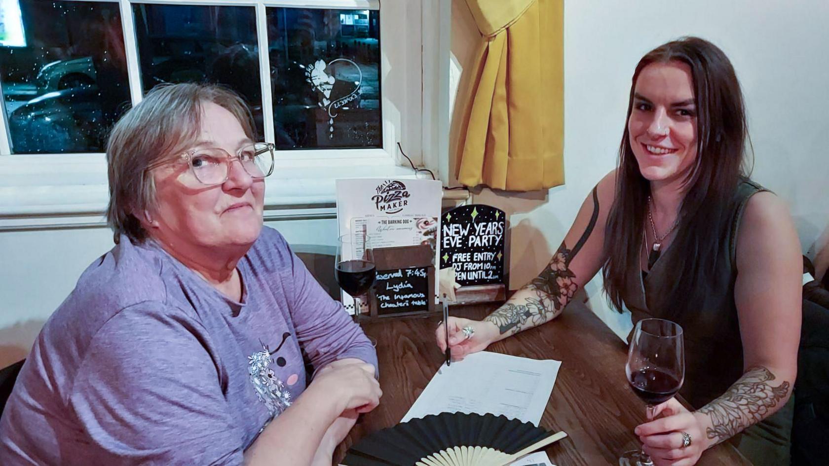 Lynda (left) with short grey hair wearing a lilac t-shirt and glasses with her daughter Lydia with long dark hair wearing a black sleeveless top sitting at a table in a pub by the window with a drink and pen in hand.