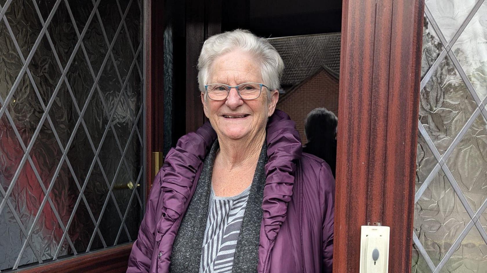 Ruth Pressley is pictured in a doorway. She wears a purple coat and glasses.