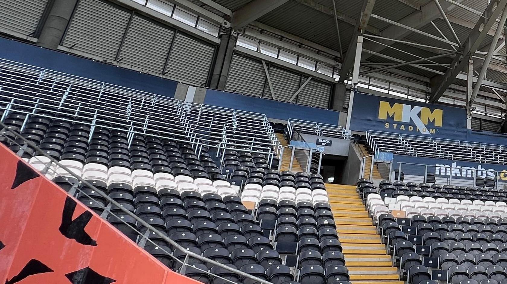 An empty sport stadium stand with rows of black and white seats. Yellow steps form a walkway between blocks. Advertising hoardings are visible. To the left, a wall has been painted orange and black to resemble tiger stripes.