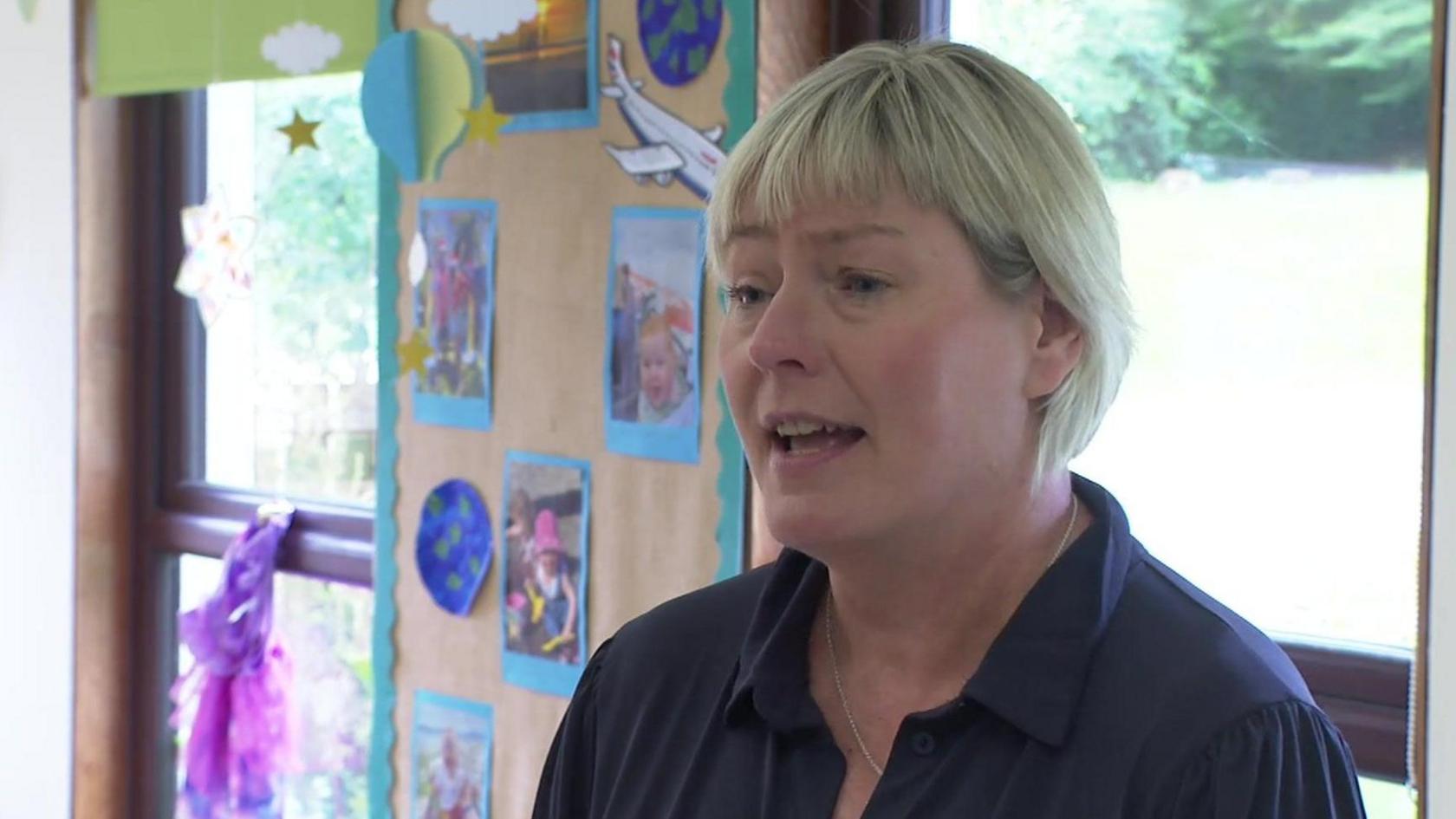 Carol Shaw has short blond hair and is wearing a purple shirt. She's standing in a classroom with pupil's artwork pinned on the wall.