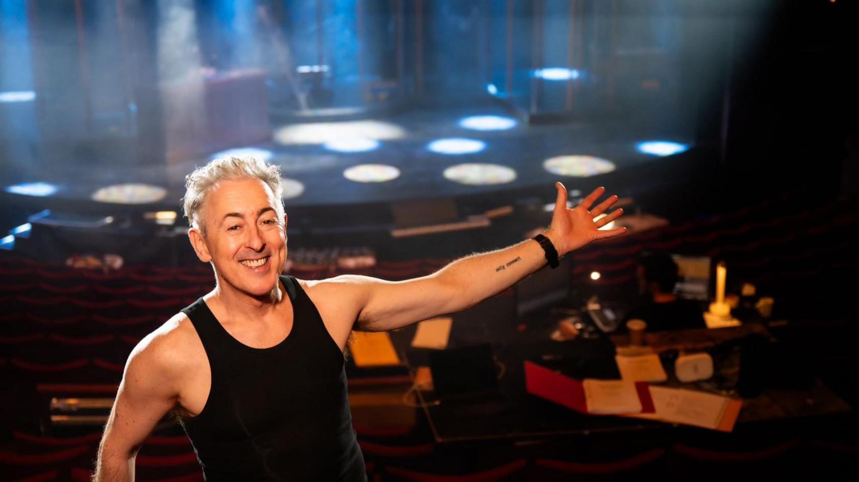 Alan Cumming standing in a theatre with an arm gesture that suggests he's showing people around. He is wearing a black vest top