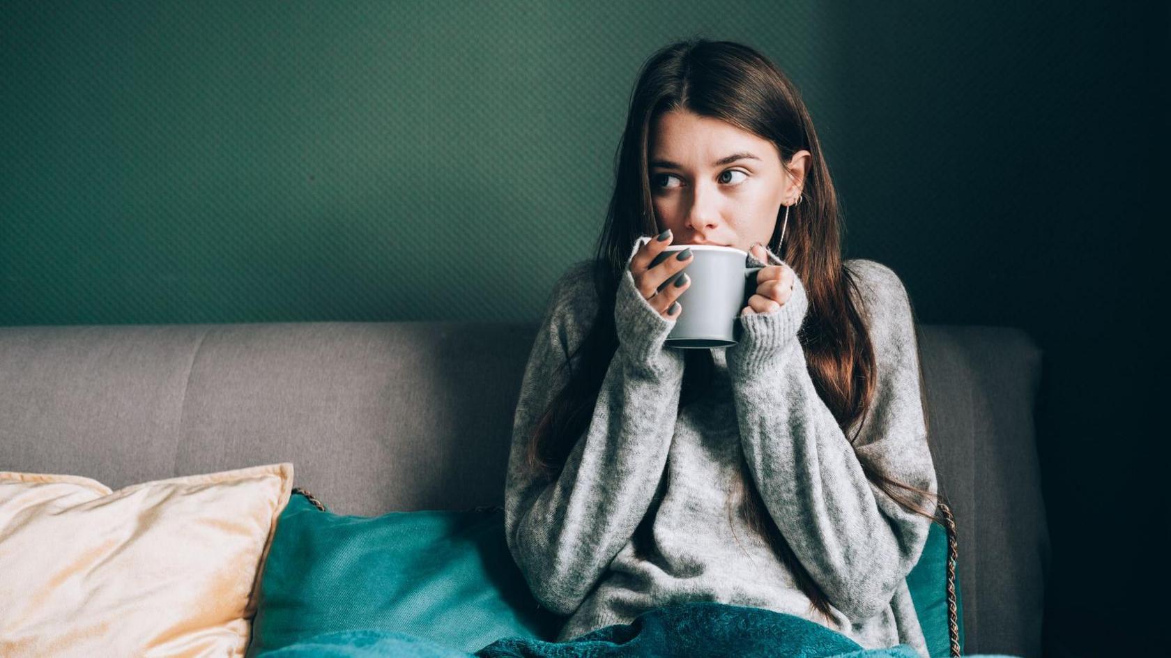Stock photo shows a woman in a jumper under a blanket with a mug of tea. She is hunching her shoulders as if feeling cold while sitting on the sofa at home.