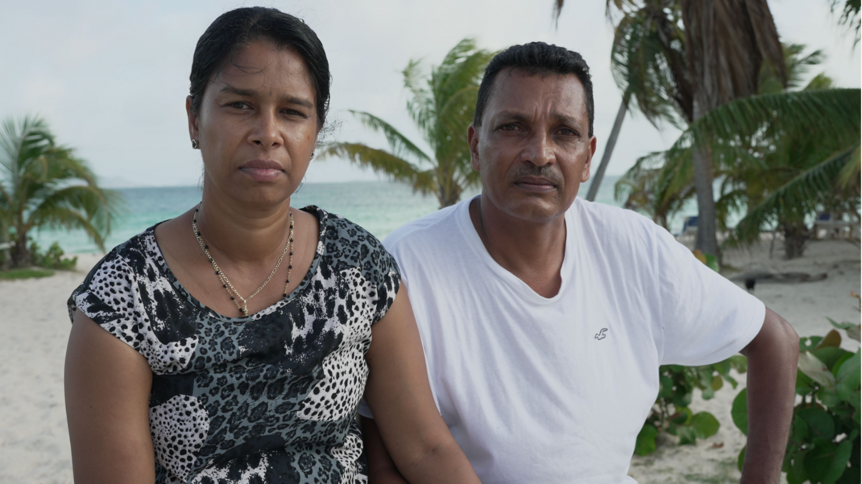 A woman and a man sit on a beach, with sand, sea and palm trees in background. The woman is wearing a black and white top. The man is wearing a white t-shirt.