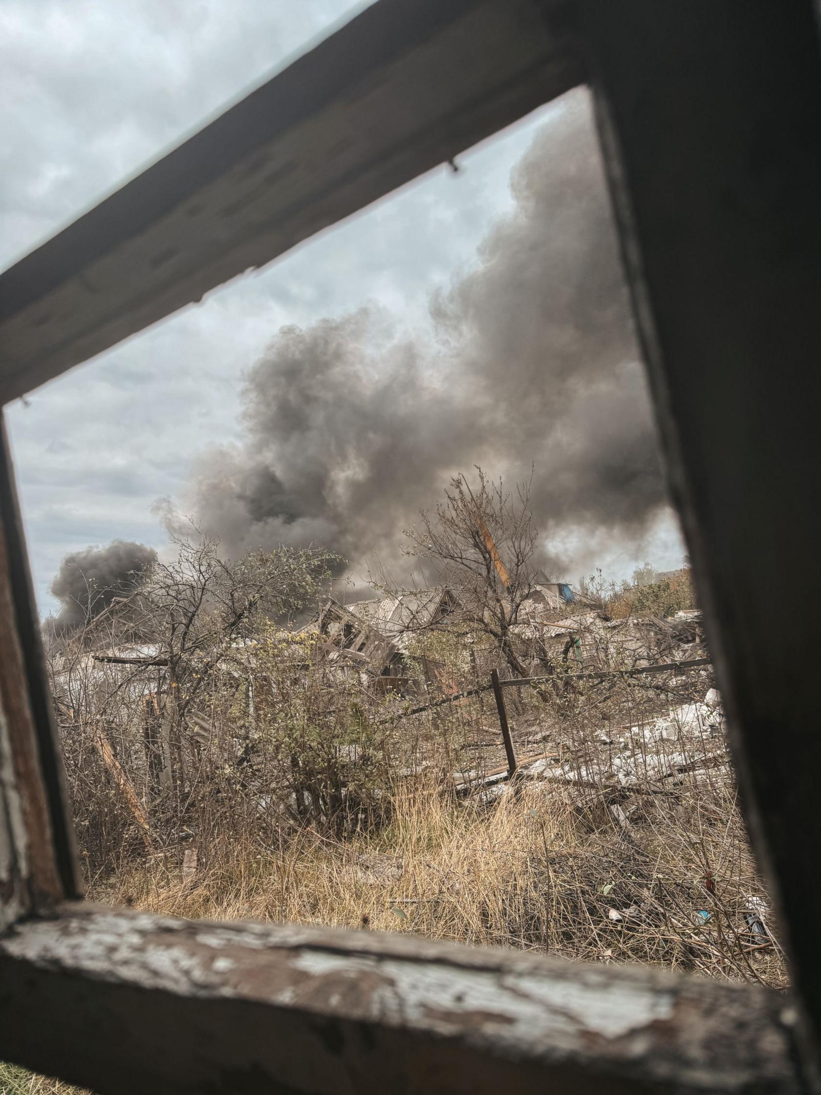 A shot through a window of a wasteland in Pokrovsk. The houses are severely damaged, the trees are bare and there is a large grey-black cloud in the distance