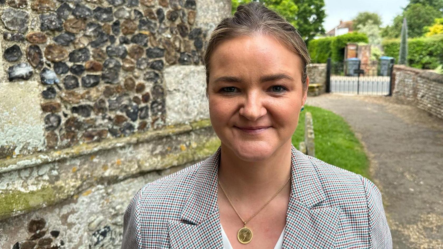 Jenny Riddell-Carpenter MP wearing a checked jacket and a white top stands in front of a church. She has brown hair, which is tied back, and is wearing a gold pendant. She is smiling at the camera.