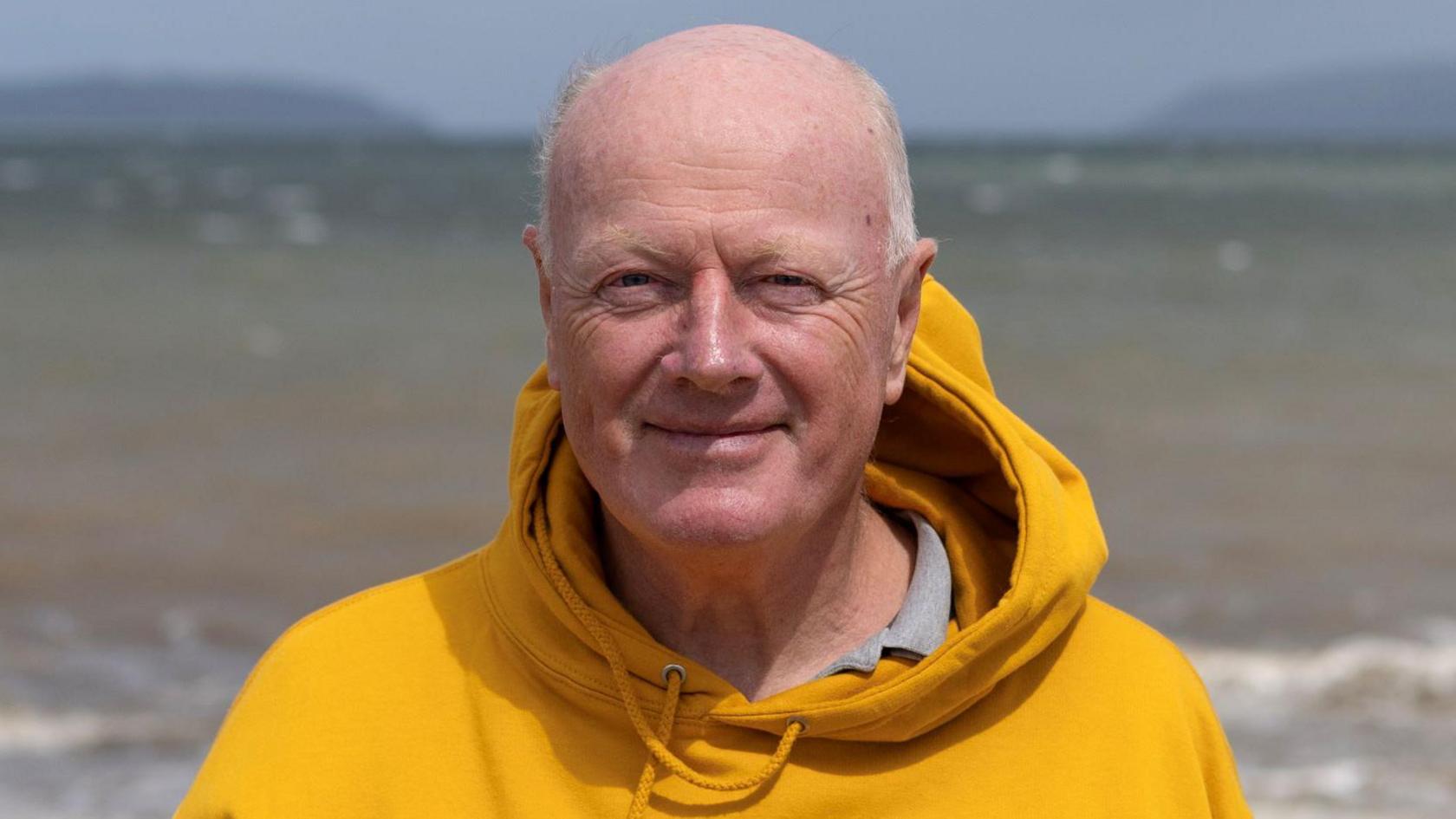 Topher is smiling as he looks into the camera. He is balding with grey hair around the sides of his head. He is wearing a bright, mustard-coloured hooded top over a grey shirt. Behind him is the sea. It is a sunny day.