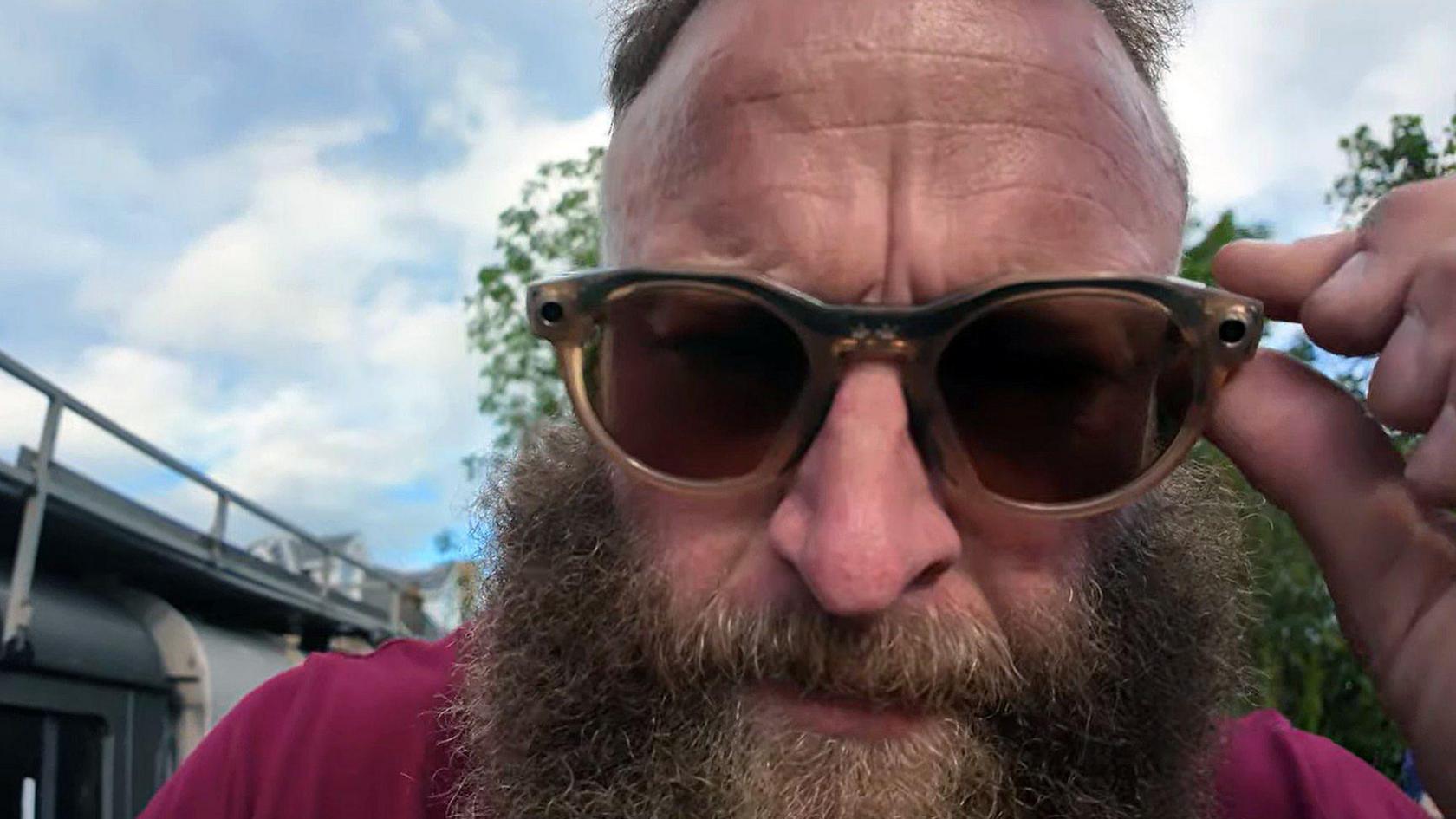 A bearded man in a red T shirt wearing sunglasses looks at the camera