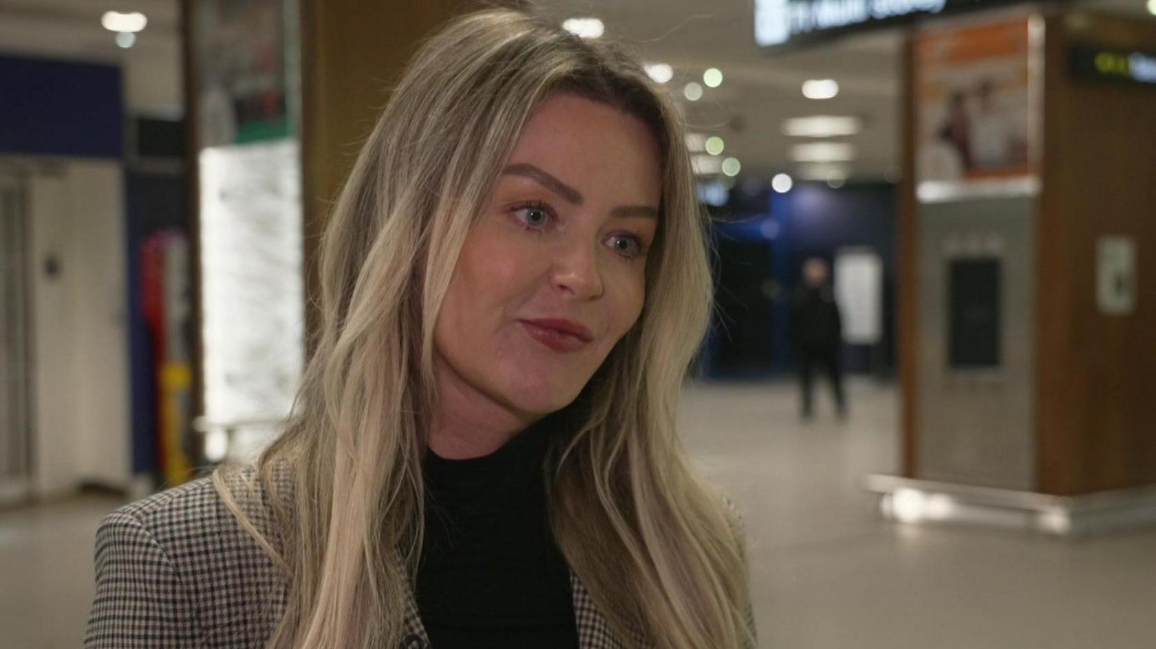 Jennifer Byrne-Smith stands in an airport terminal, she has long blonde hair and wears a tweed blazer over a black roll-neck jumper. She is looking to the right. 