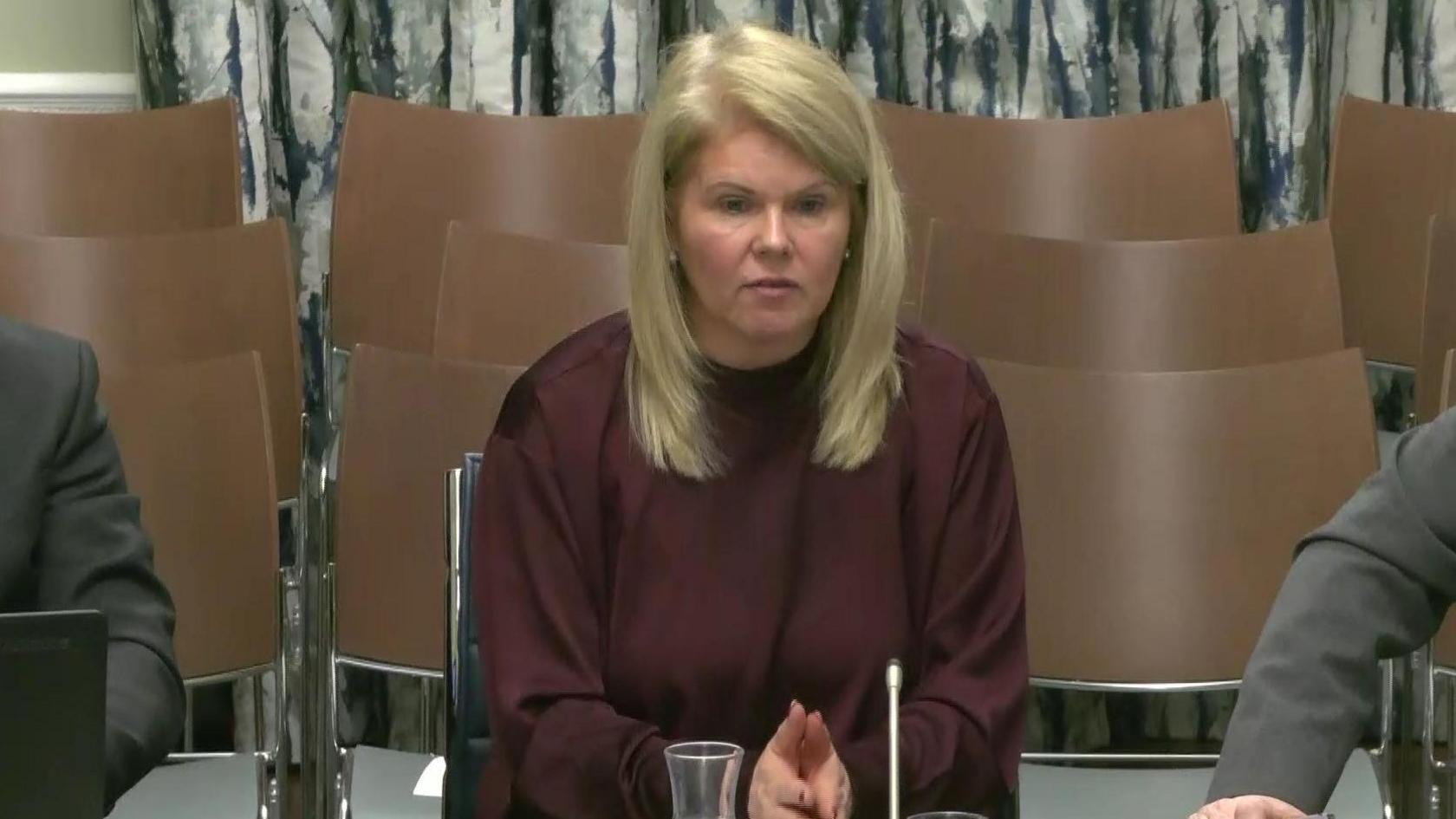 NI Water Chief Executive Sara Venning answering questions from assembly members at Stormont's infrastructure committee.   She has straight, shoulder-length blonde hair and is wearing a loose-fitting maroon crew-neck top or dress.