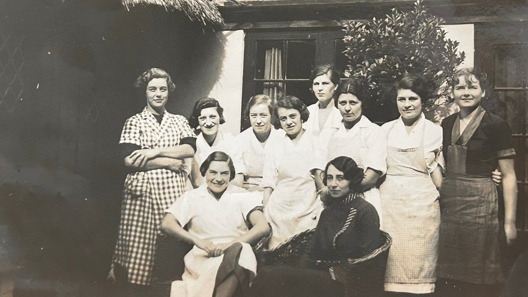 Ten women stood in front of a building. The woman are wearing a uniform of white shirts with an apron. They are stood in three rows.