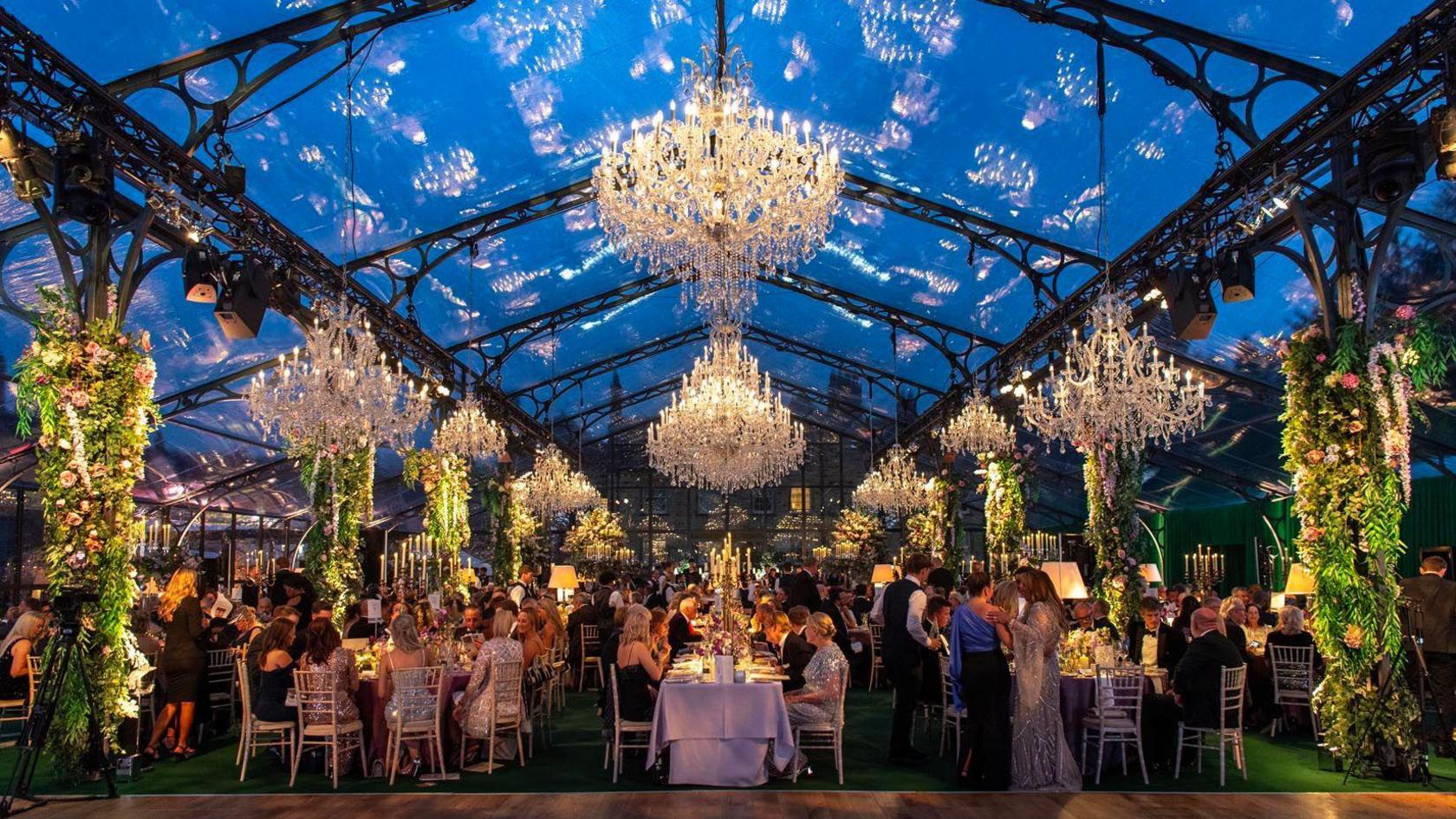 Guests sat on several long tables inside a glass orangery, with decorative flowers and chandeliers 