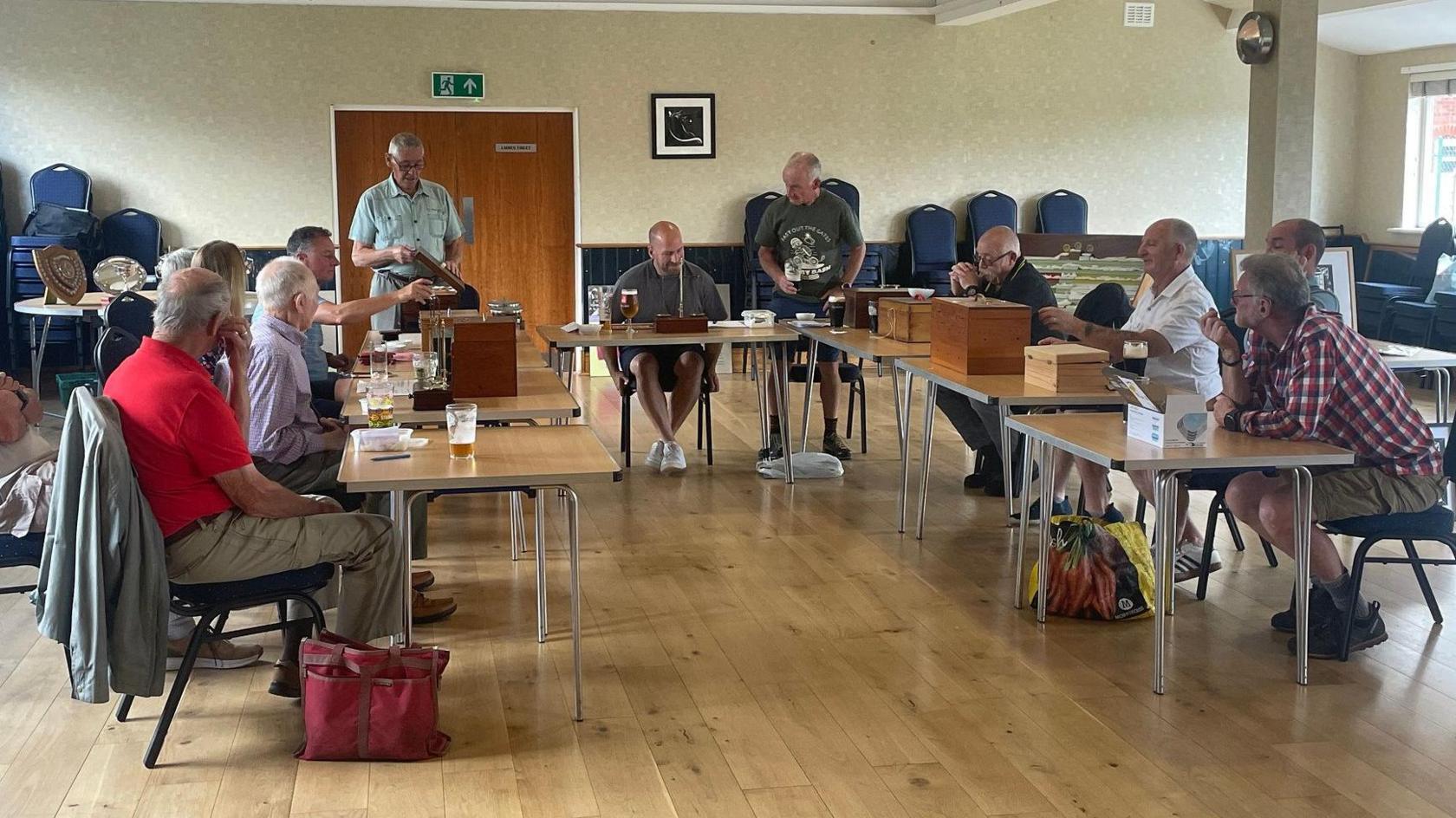 About 10 men can be seen sitting at rows of table with their produce in front as fruits get weighed or monitored. They are in a large room inside a centre with chairs stacked around.