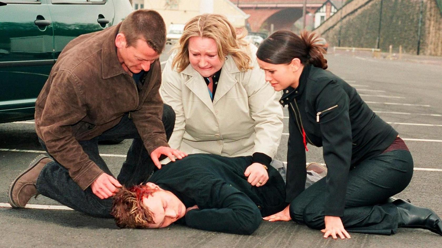 Annie Orwin as Lou Gallagher in Byker Grove alongside Andrew Hayden Smith as Ben and Holly Matthews as Emma. In the scene, she and Emma crouch over Ben who's just been hit by a green car, his head bleeding as he lies face down in a car park.