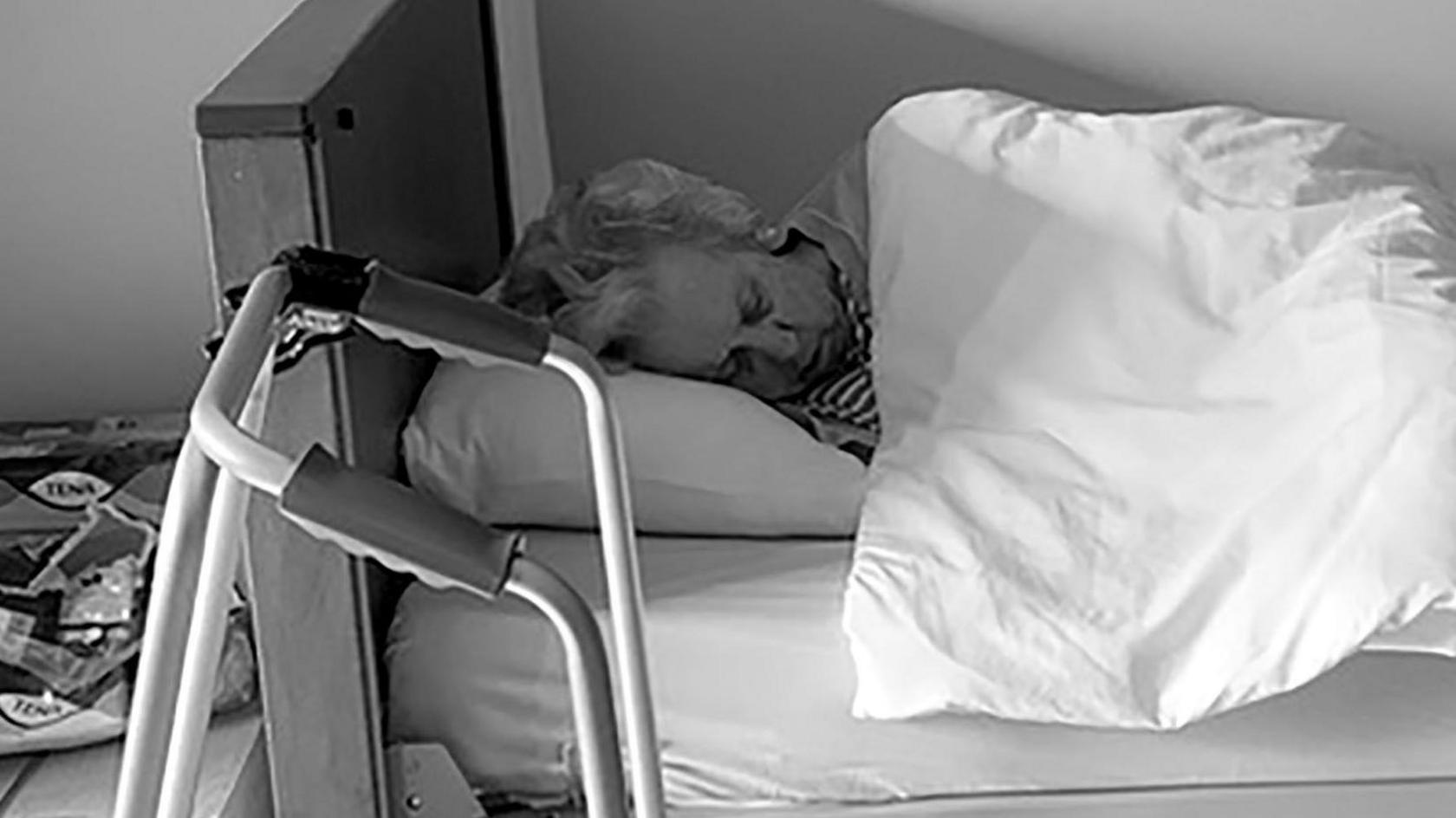Jenny Harris's mother asleep in a hospital bed in a care home with a wheeled zimmer frame close to it